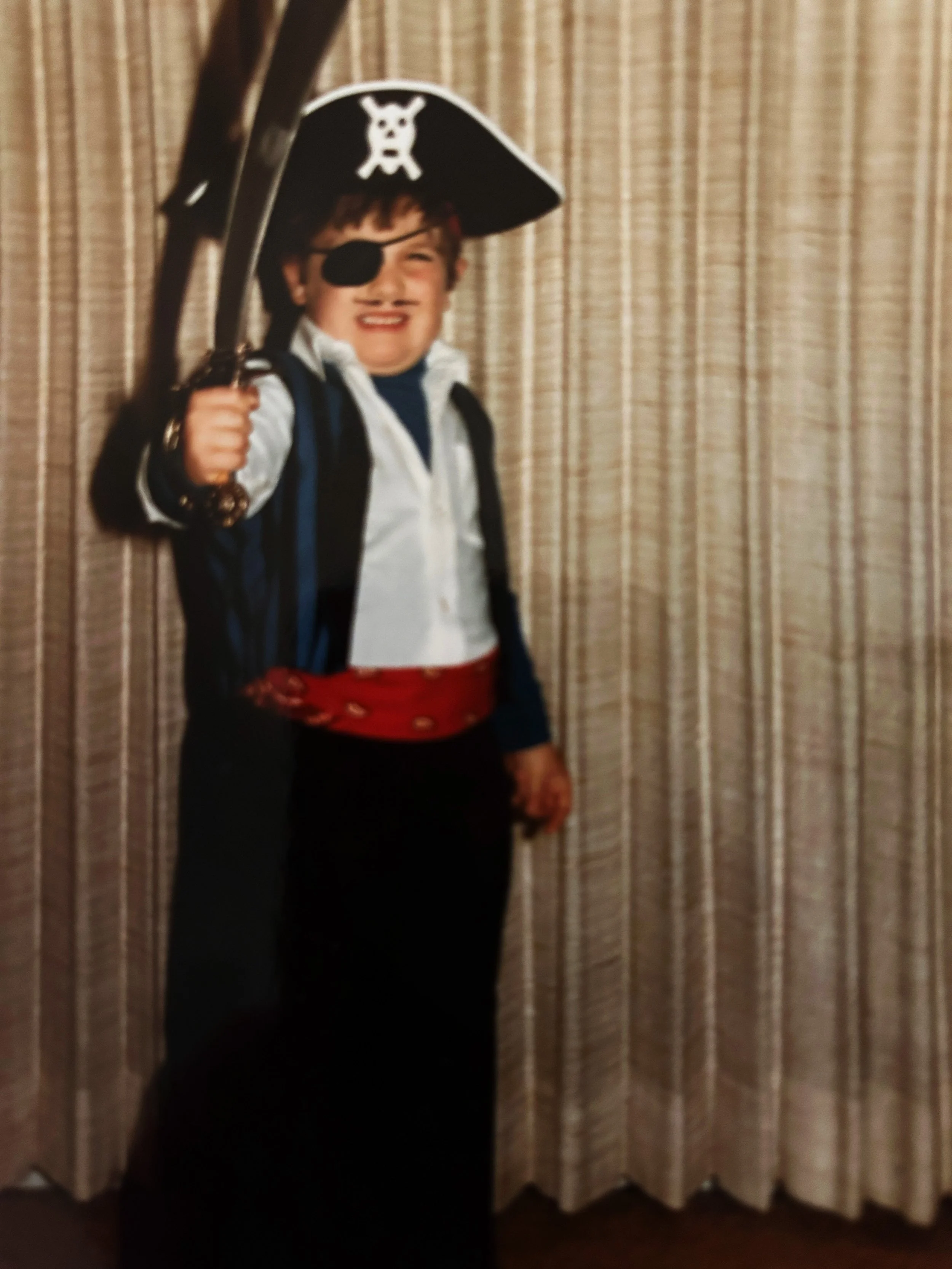 Child dressed as a pirate with an eyepatch, a pirate hat with a skull and crossbones, holding a toy sword, standing in front of beige curtains.