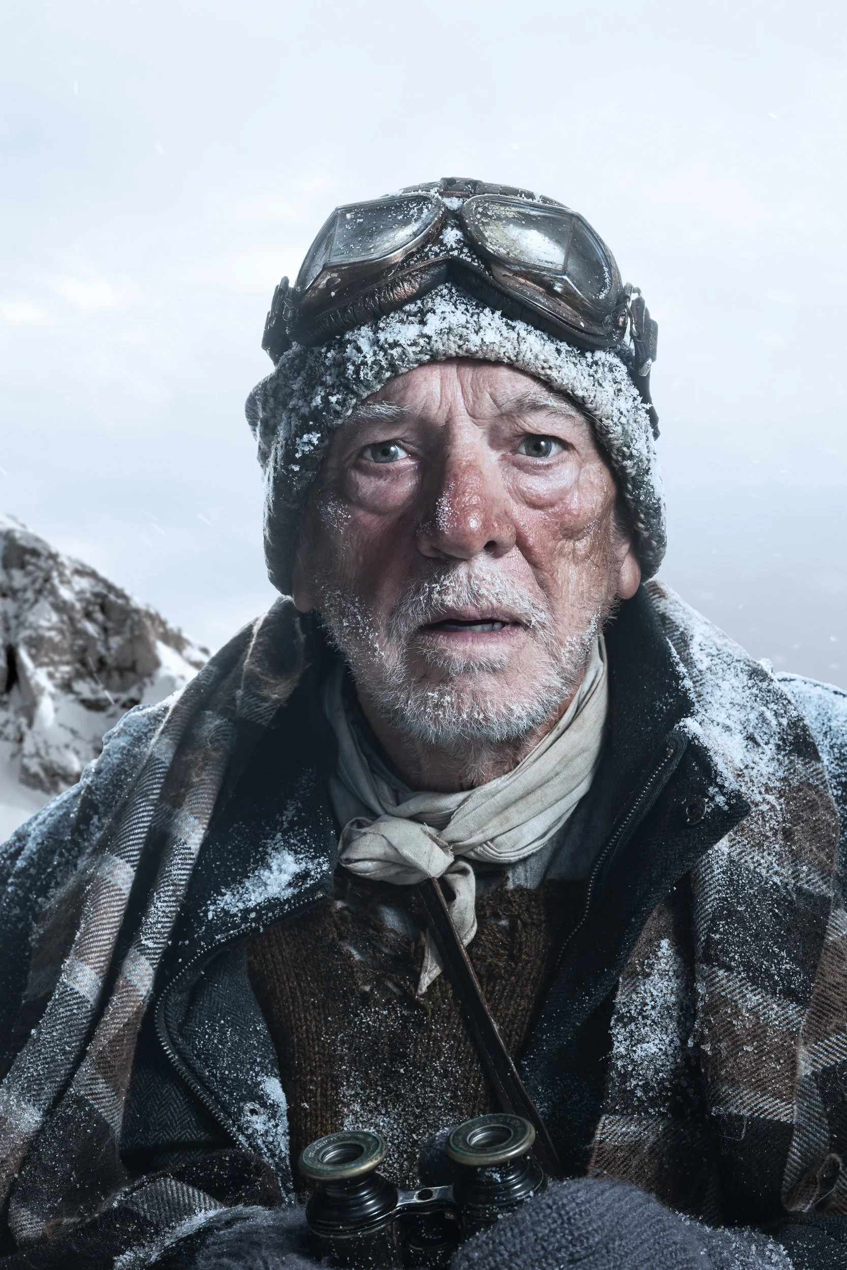 A snowy mountain climber with goggles on his head, wearing a beanie and winter clothing, holding binoculars, surrounded by snow and overcast sky.