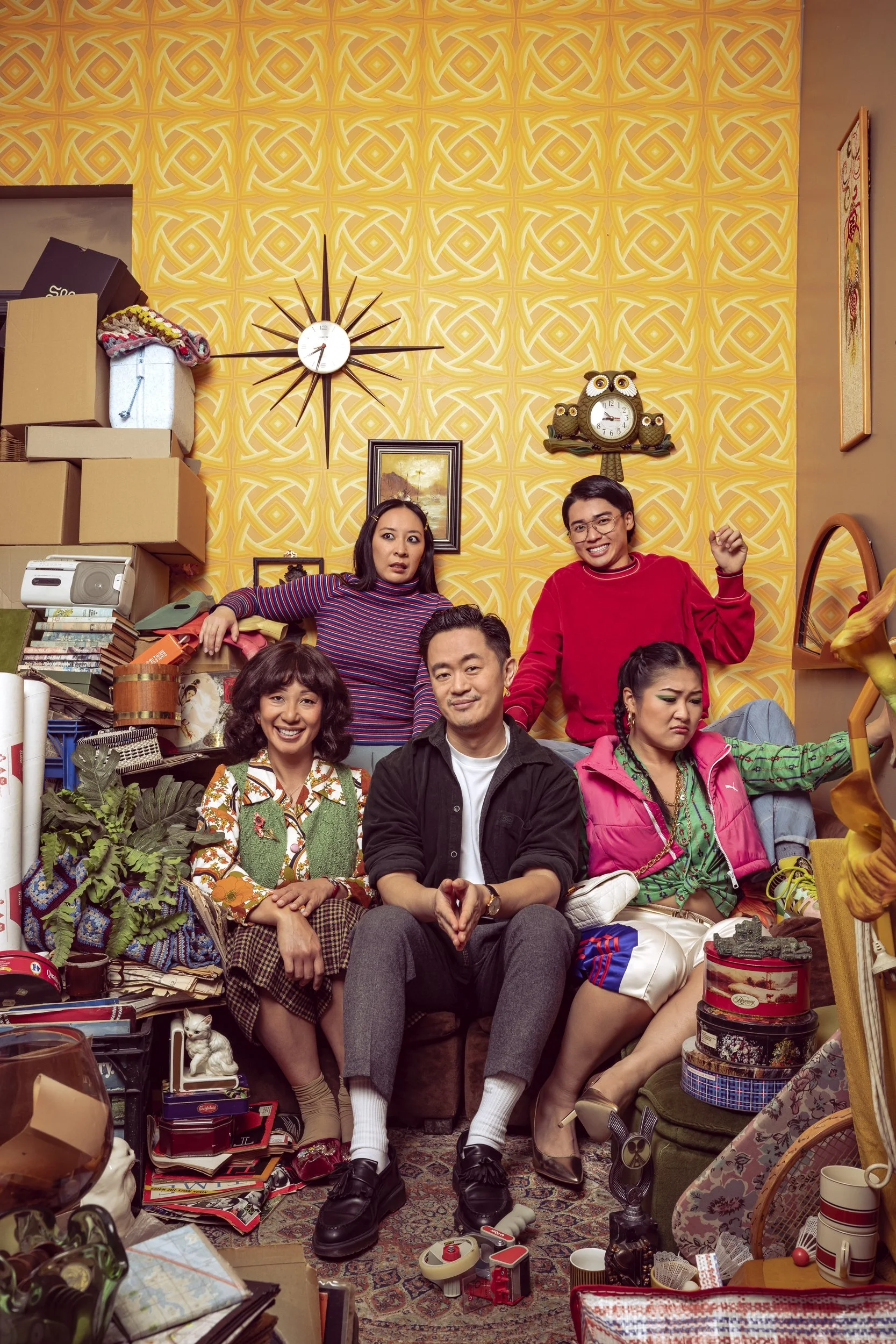 A diverse group of people in a cluttered room with yellow patterned wallpaper, various objects, paintings, clocks, and boxes. They are sitting and standing, some smiling and some looking confused.