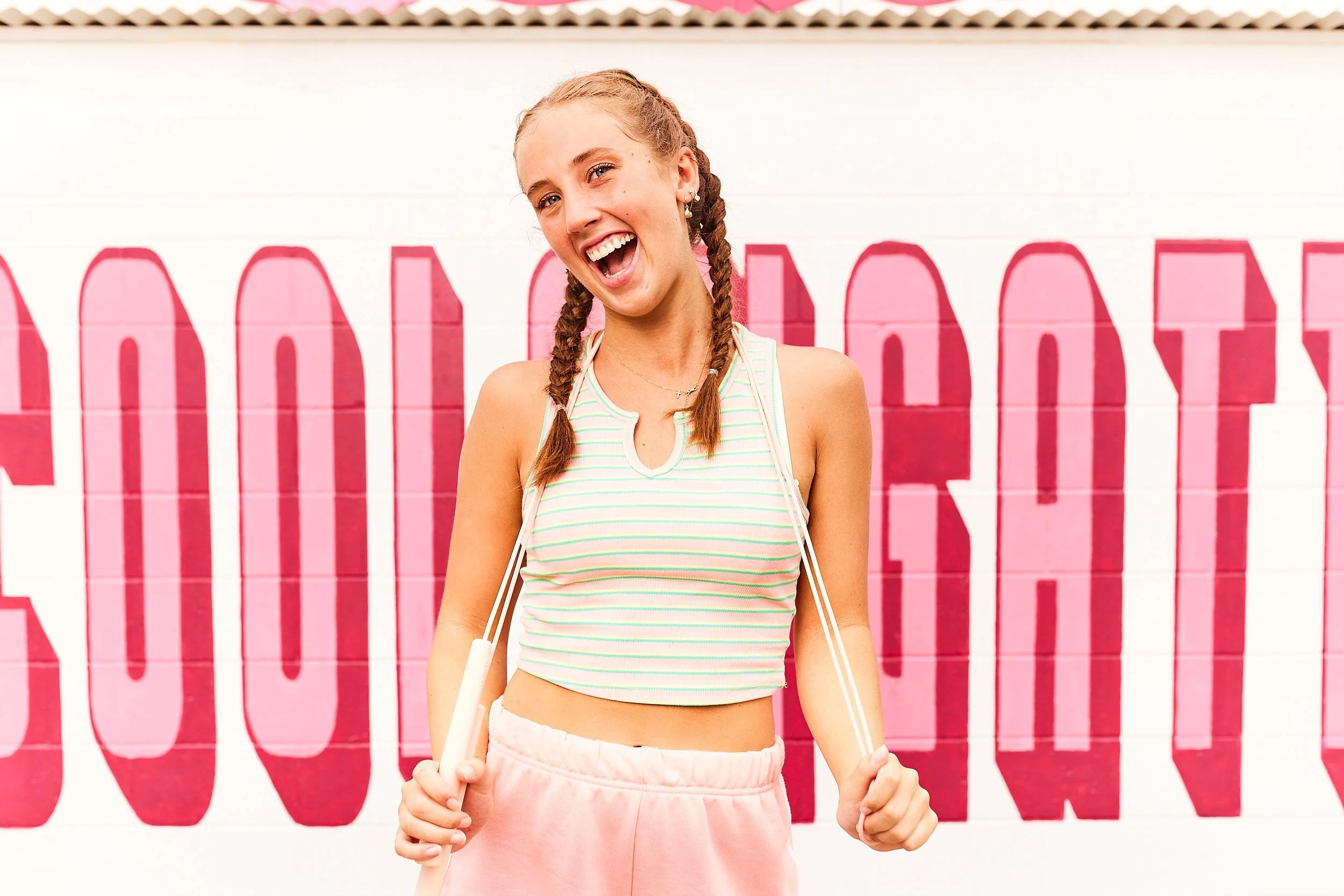 A young woman with braided hair, wearing a striped tank top and pink shorts, smiling and holding the straps of her bag in front of a pink and white wall with bold lettering.