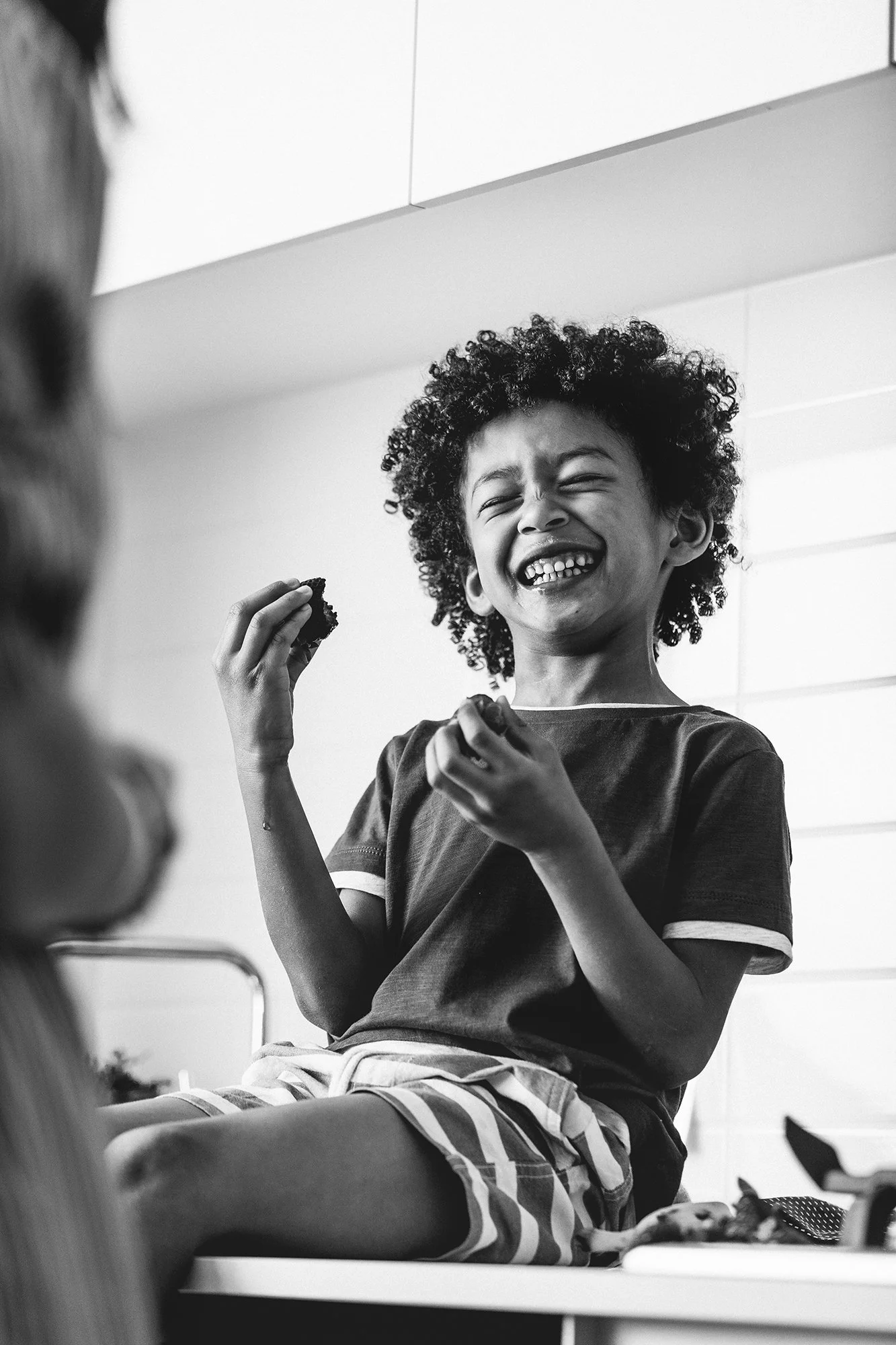 A young boy with curly hair smiling and laughing while holding a snack in his right hand in a classroom or indoor setting.