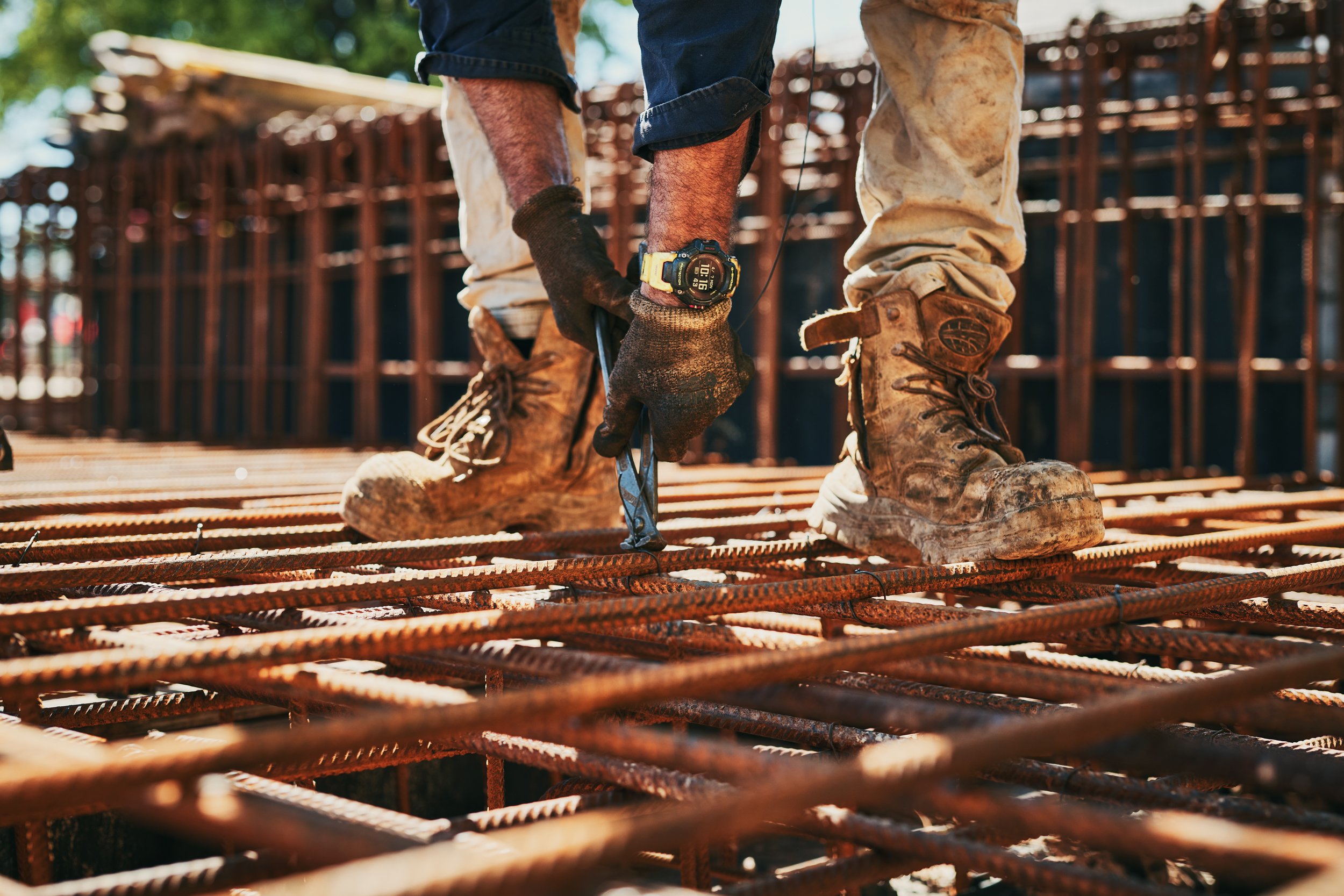 Close-up of a construction worker's legs and hands as he works on a rebar foundation at a building site, wearing work boots, gloves, and a yellow watch.