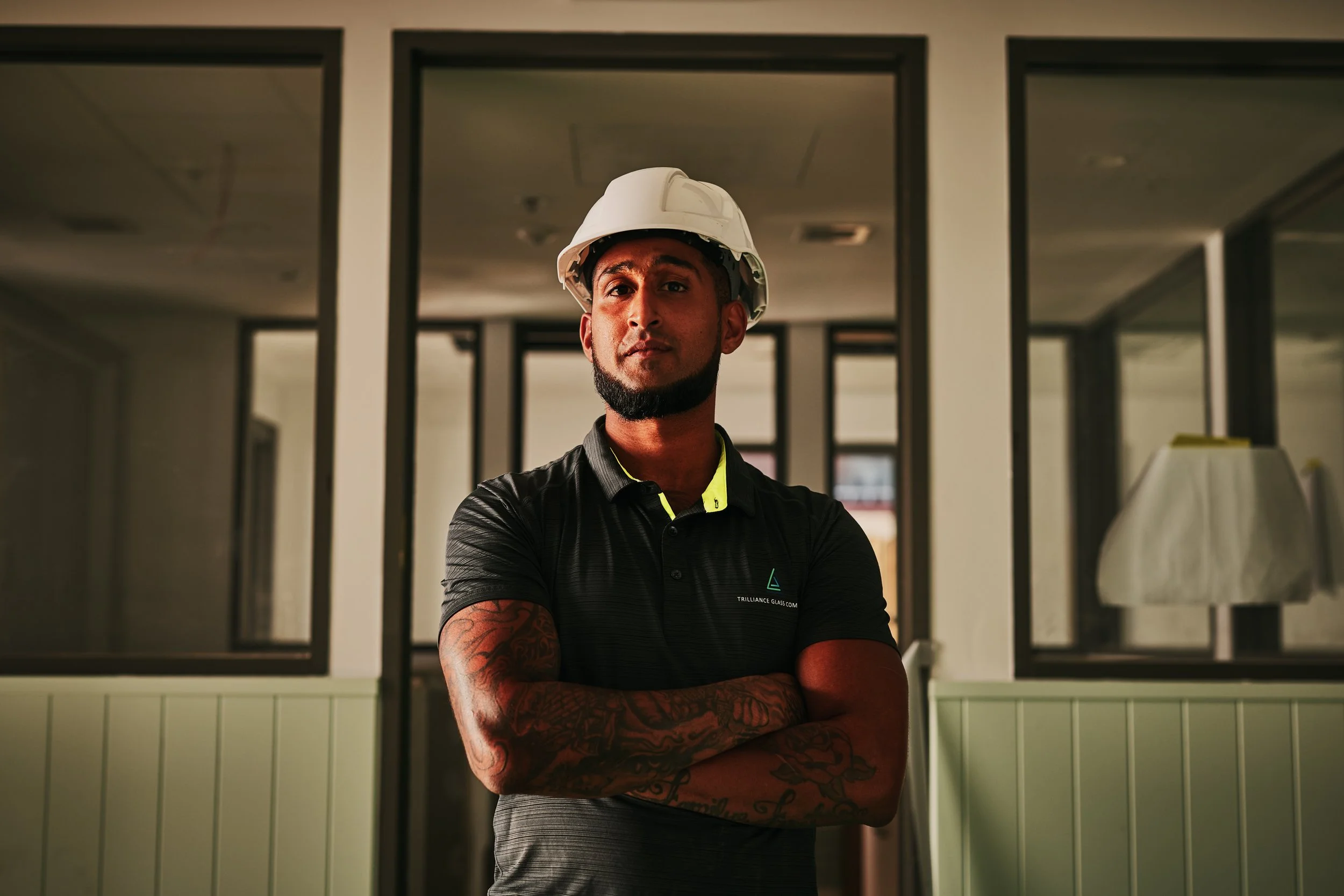 A man with tattoos on his arms wearing a white safety helmet and a black polo shirt with a logo stands with his arms crossed in front of a windowed wall inside a building.