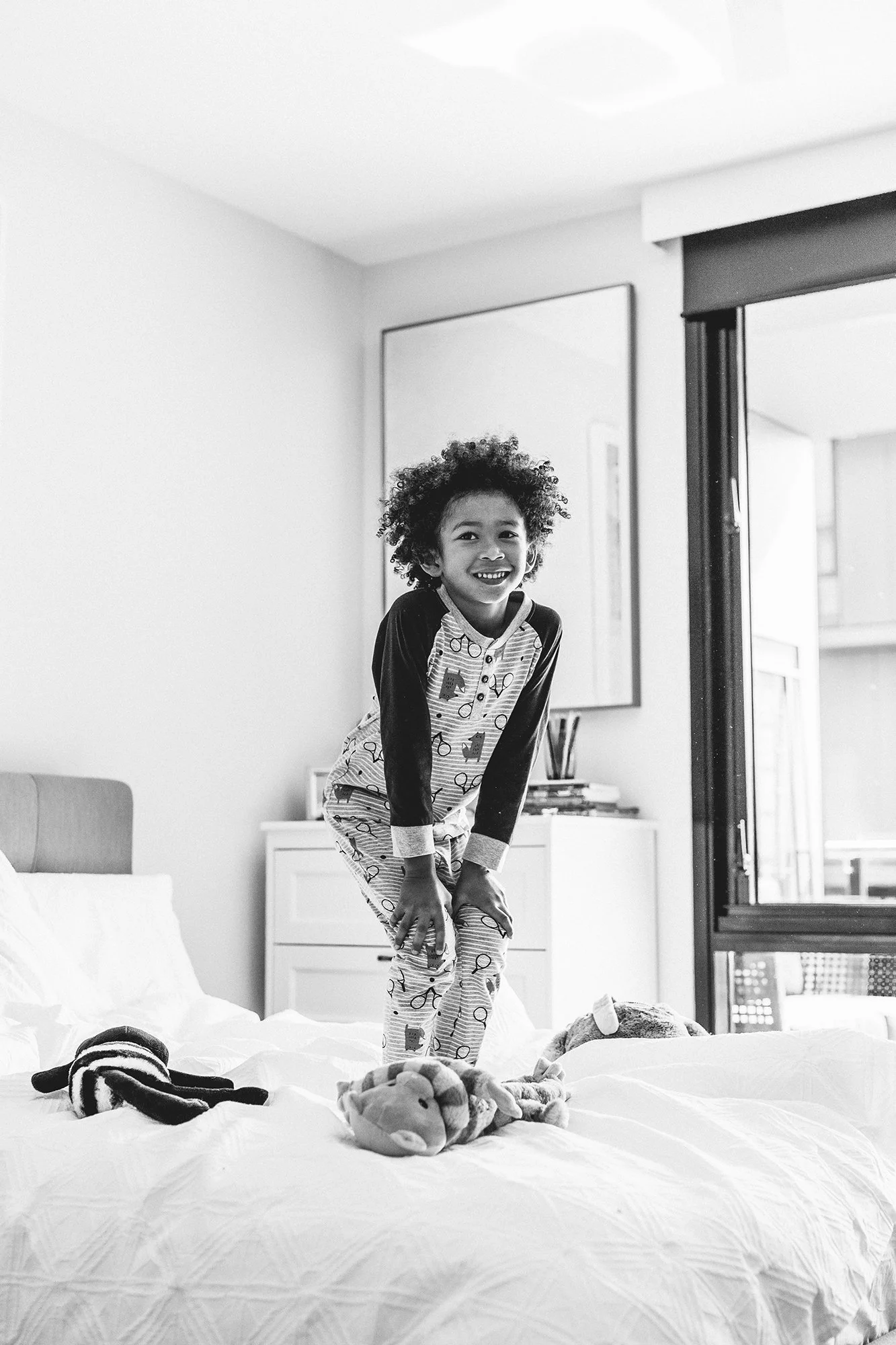 A smiling child with curly hair in pajamas, standing on a bed with stuffed animals in a bedroom, near an open sliding glass door.