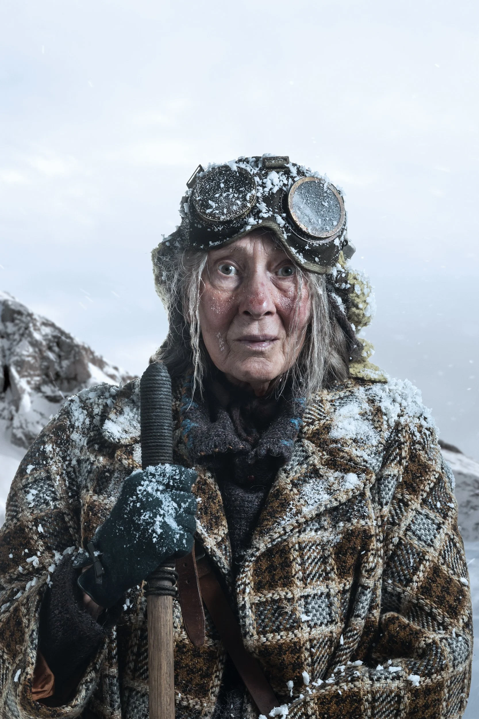 Close-up of an elderly person with snow-covered face and long hair, wearing a plaid coat, black gloves, and vintage goggles with snow on them, holding a wooden staff in a snowy landscape.