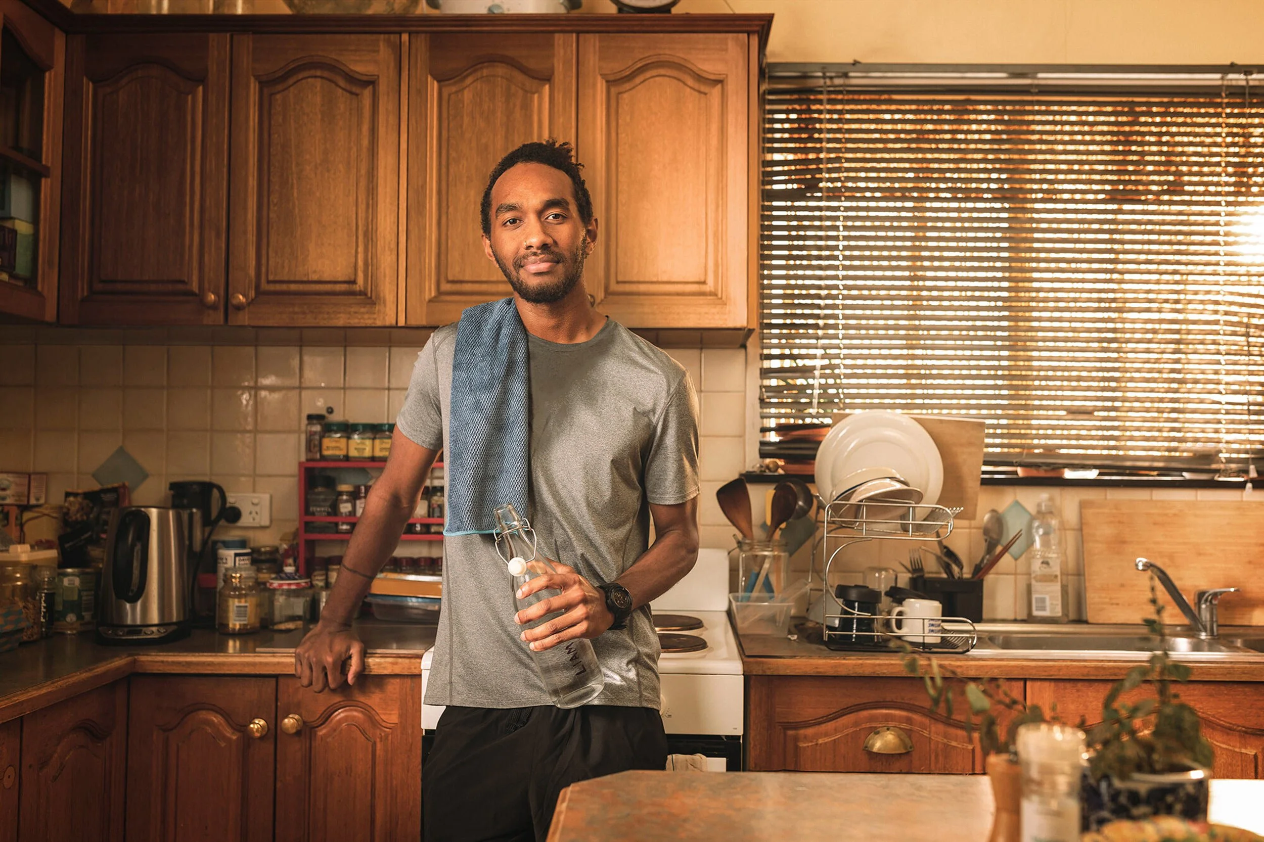 A man standing in a kitchen, holding a glass bottle, with wooden cabinets, a dish rack, and sunlight coming through window blinds in the background.