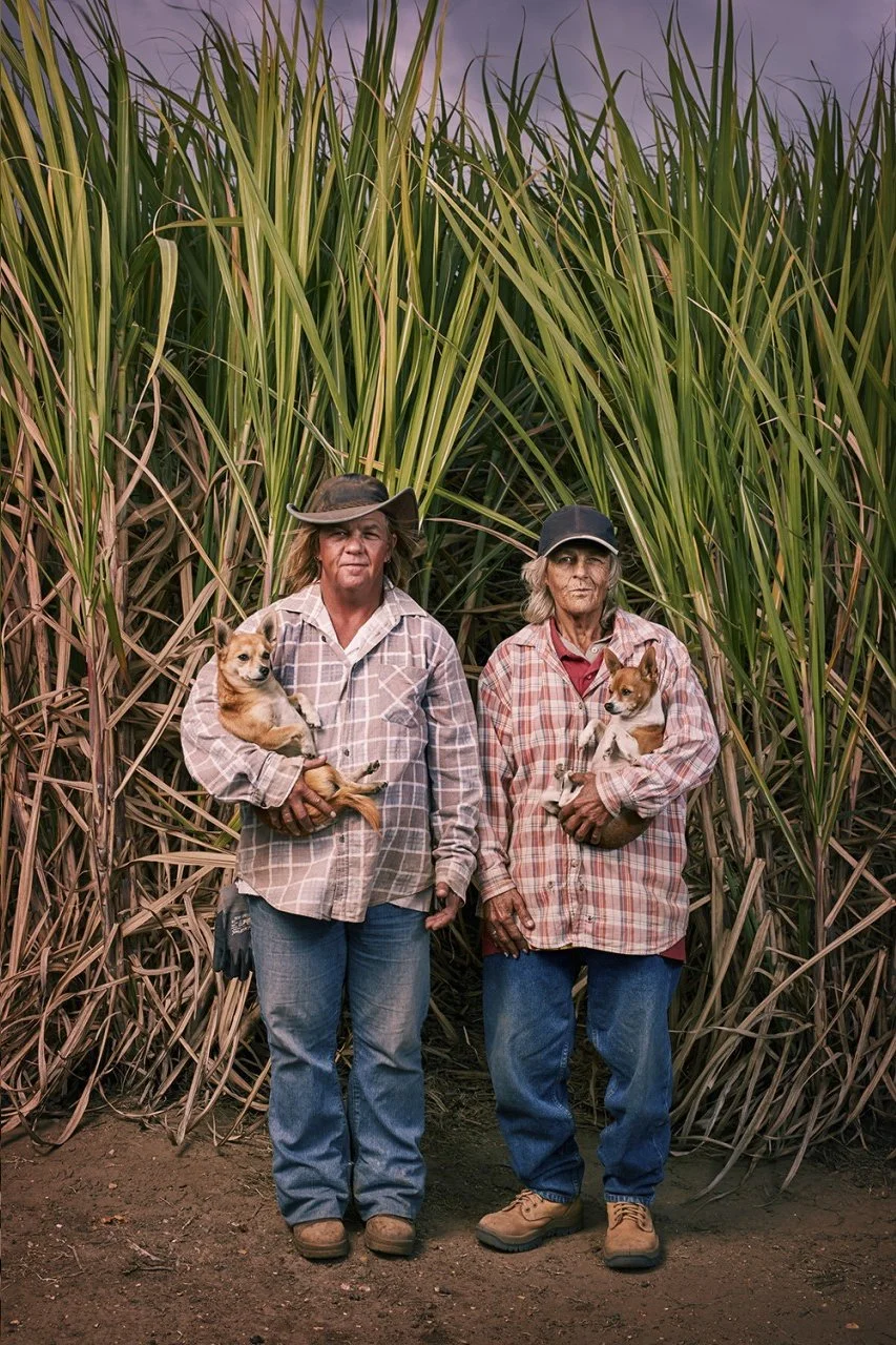 Two women standing in front of tall crop plants, each holding a small dog, dressed in casual, plaid and denim clothing.