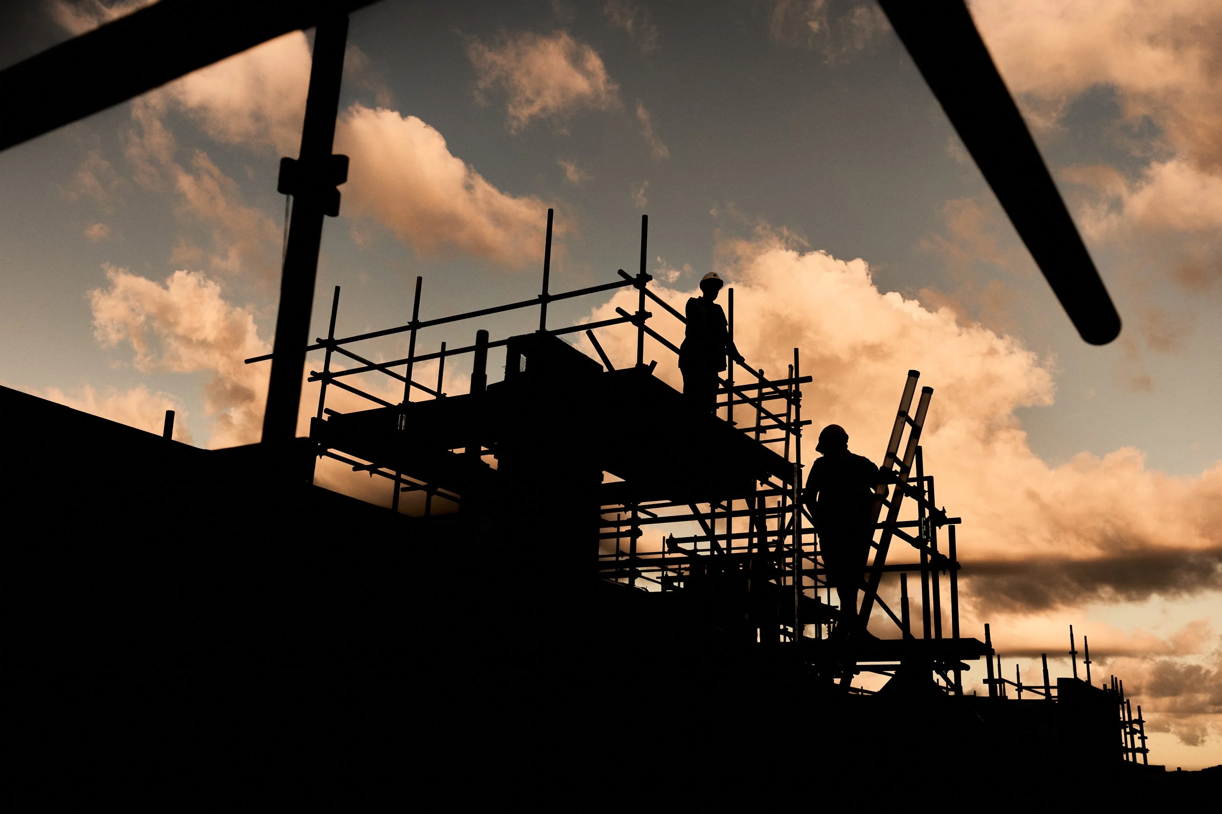 Silhouettes of construction workers on scaffolding during sunset, with a sky filled with soft, orange and pink clouds.