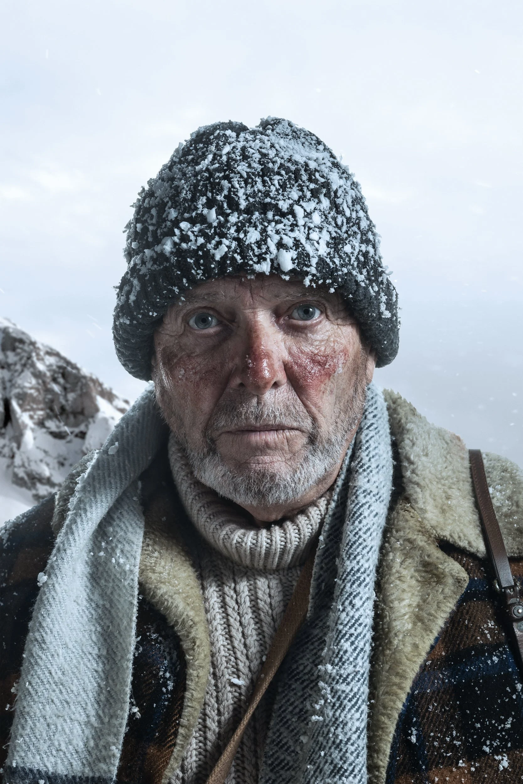Close-up of an elderly man with blue eyes wearing a thick gray knit hat covered in snow, a beige and brown plaid jacket, a beige scarf, and a cream-colored hoodie, standing outdoors in a snowy landscape during snowy weather.