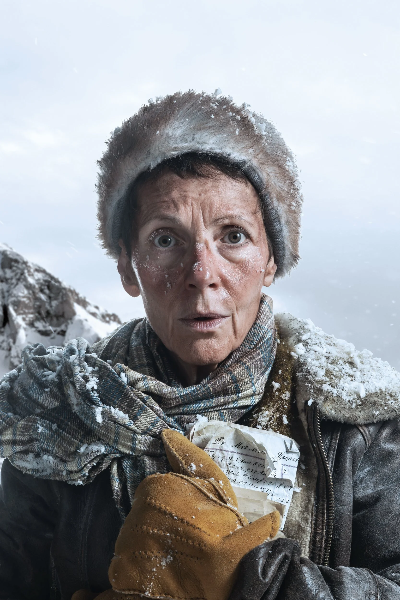 A woman with snow on her face and clothes stands outdoors in a snowy landscape, holding a piece of bread, with notes and papers in gloves in her hand, and snow on her hair and shoulders.