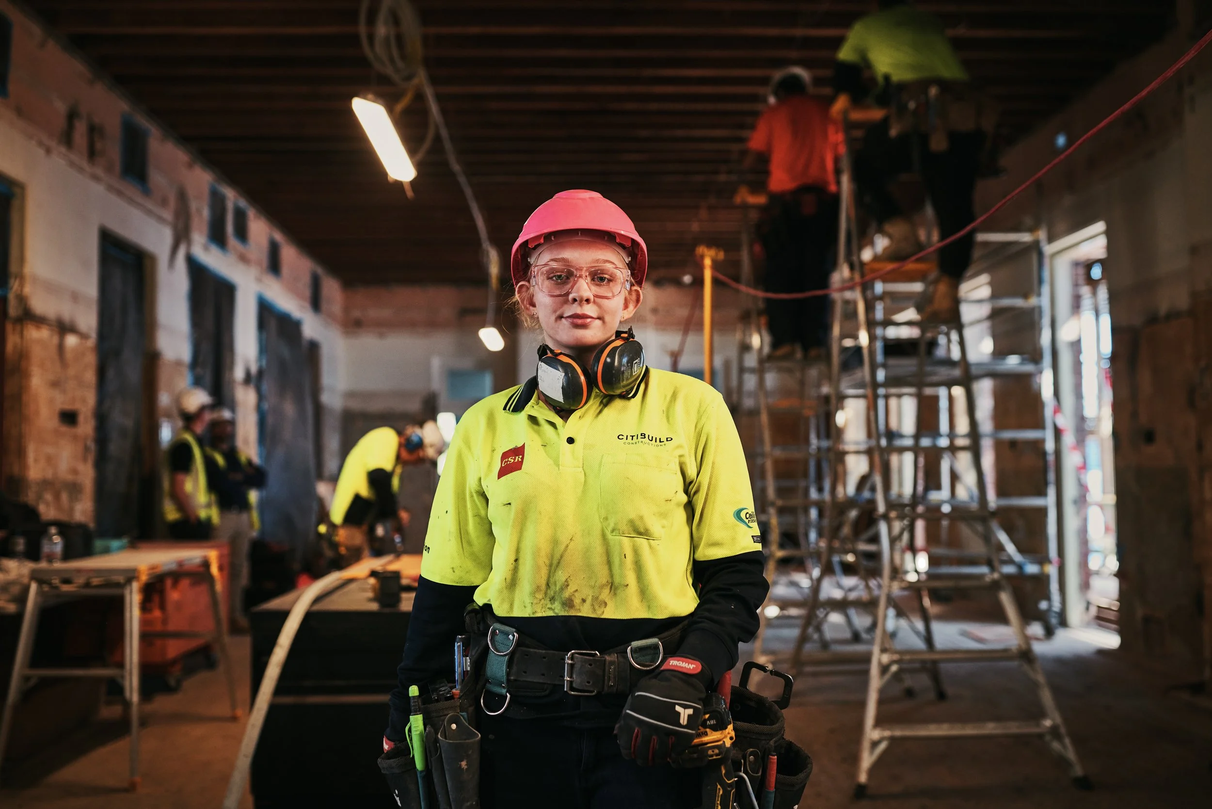 A female construction worker wearing a pink safety helmet, glasses, and a yellow safety vest standing inside a building under construction. She has a tool belt with various tools and is surrounded by scaffolding and other workers in the background.