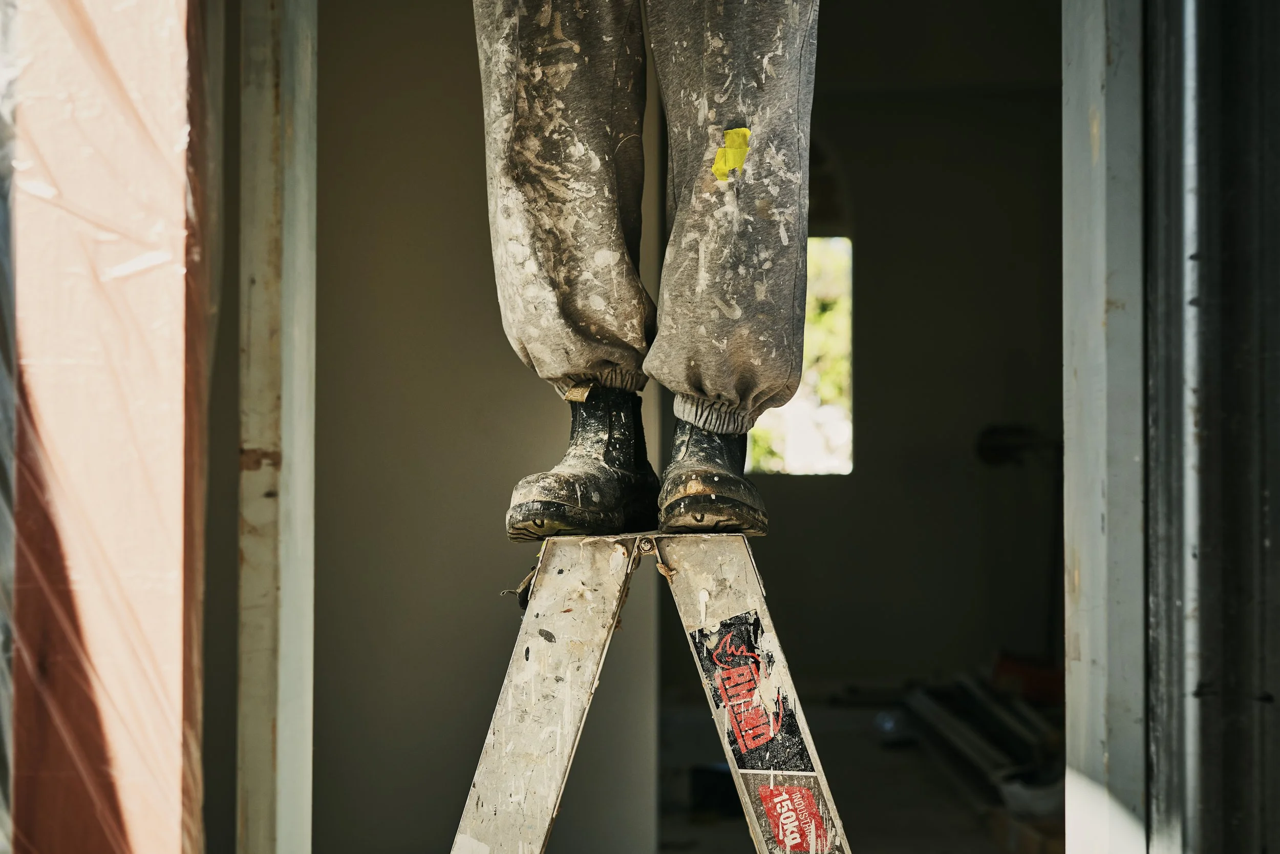 A pair of dirty work boots hanging on the top rung of a stepladder inside a building under construction, with visible wall framing and a small window in the background.