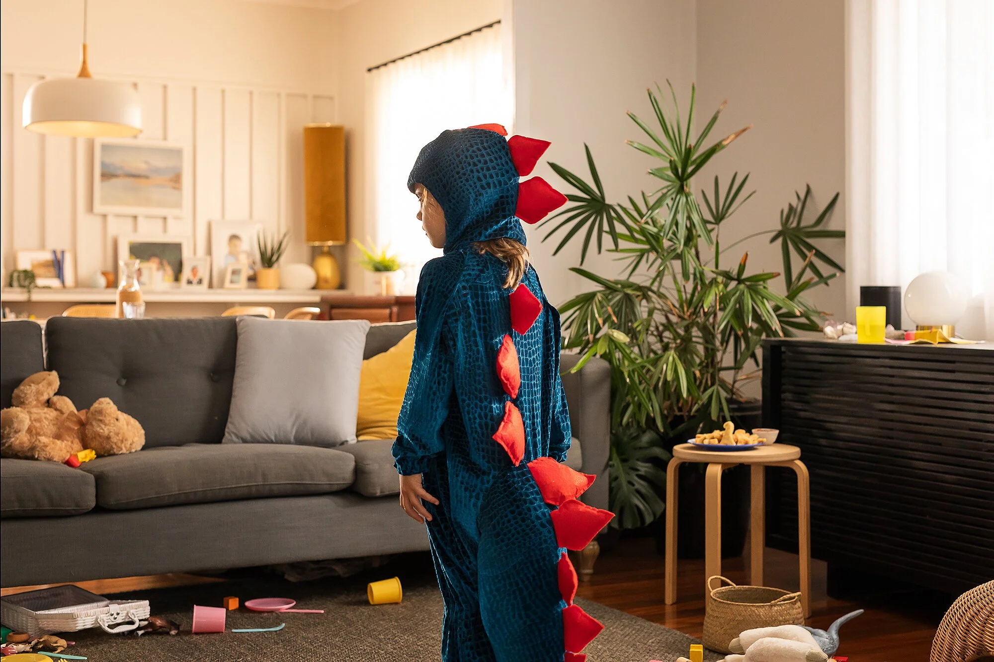 Child dressed in a dinosaur costume standing in a living room, surrounded by scattered toys and household items, with toys on the couch and a plant next to a side table.