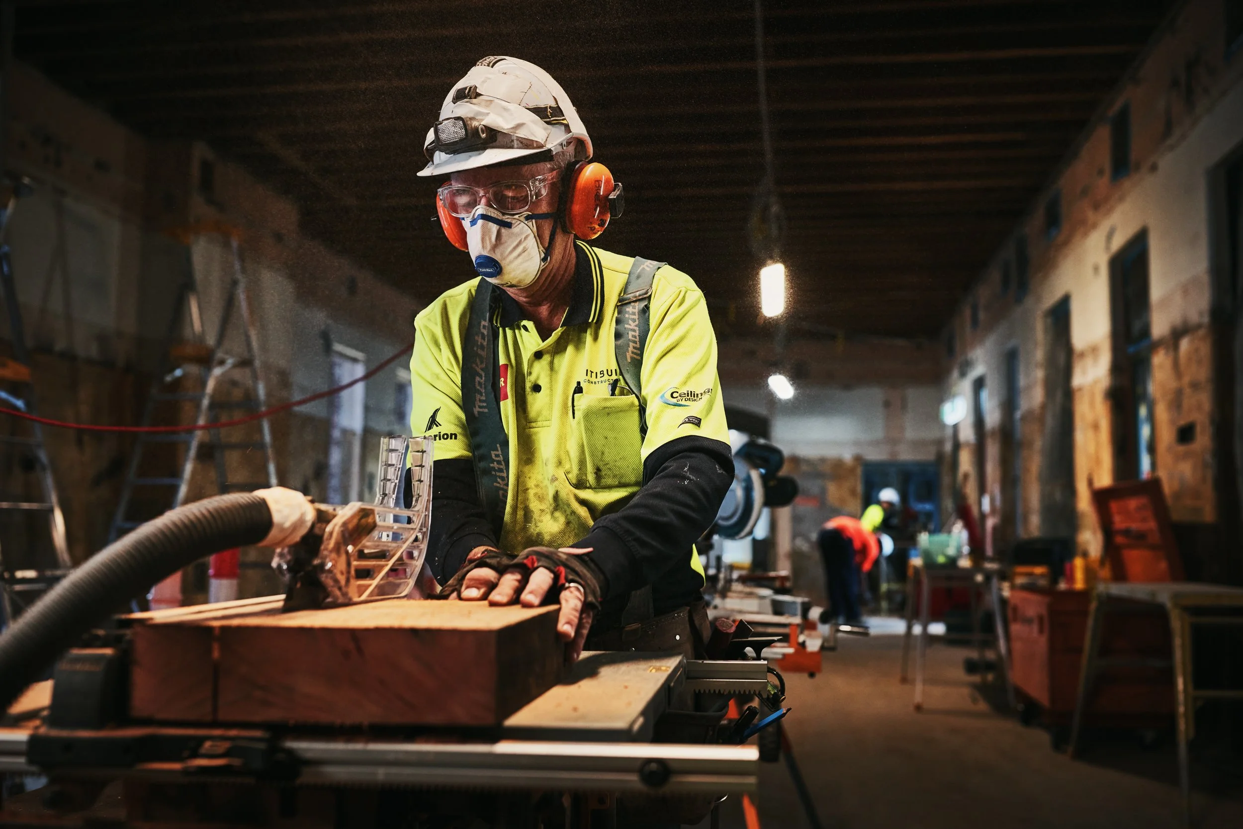 A construction worker wearing safety gear, including a helmet, goggles, ear protection, and a face mask, working on a wood project in an indoor construction site with other workers and tools in the background.