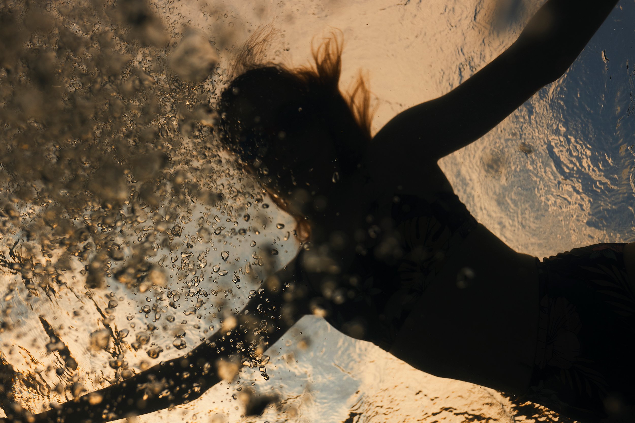 Person swimming underwater in a pool during sunset, with water bubbles around.