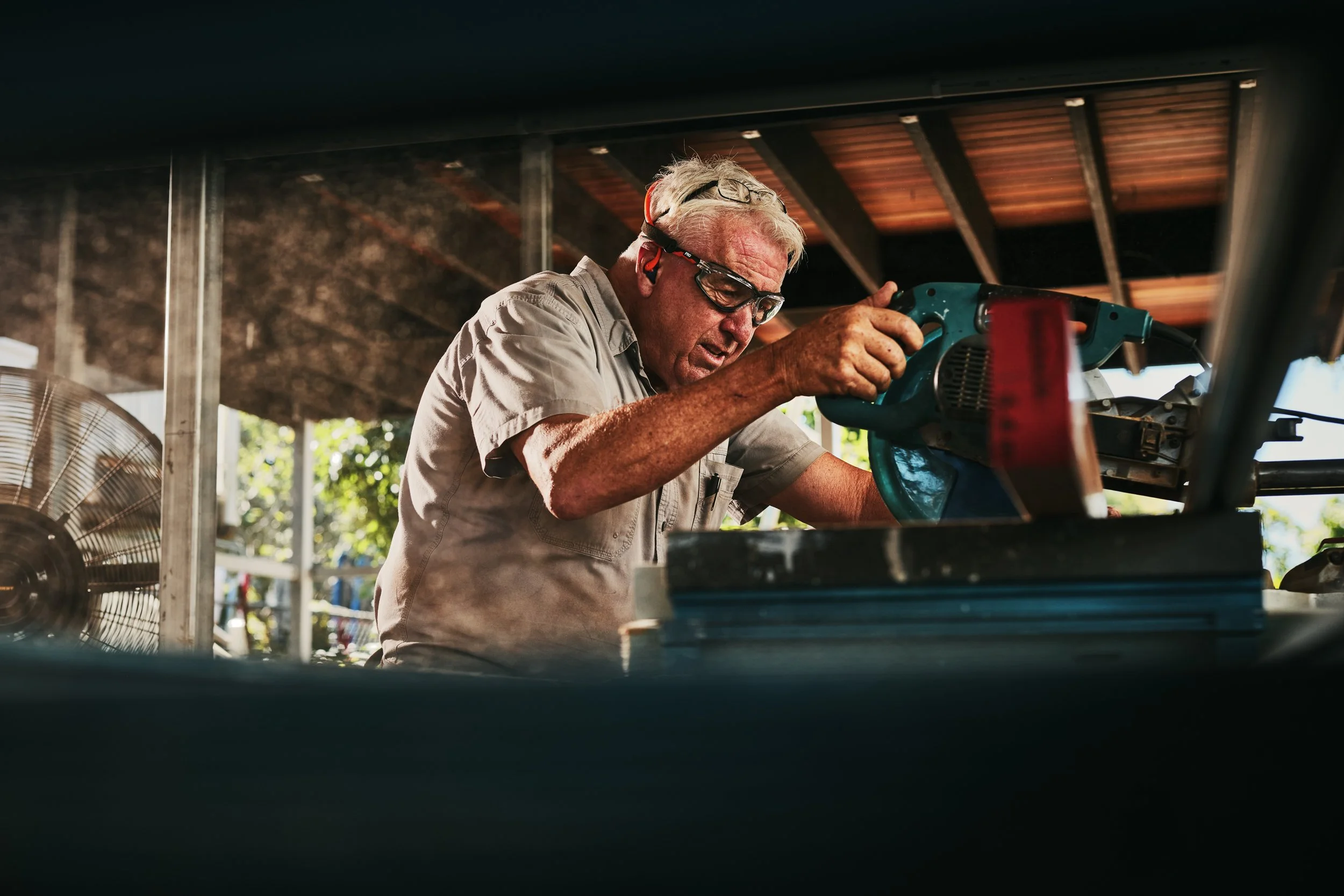 An elderly man wearing safety glasses and beige work clothes is sawing with a power saw under a roofed outdoor structure.