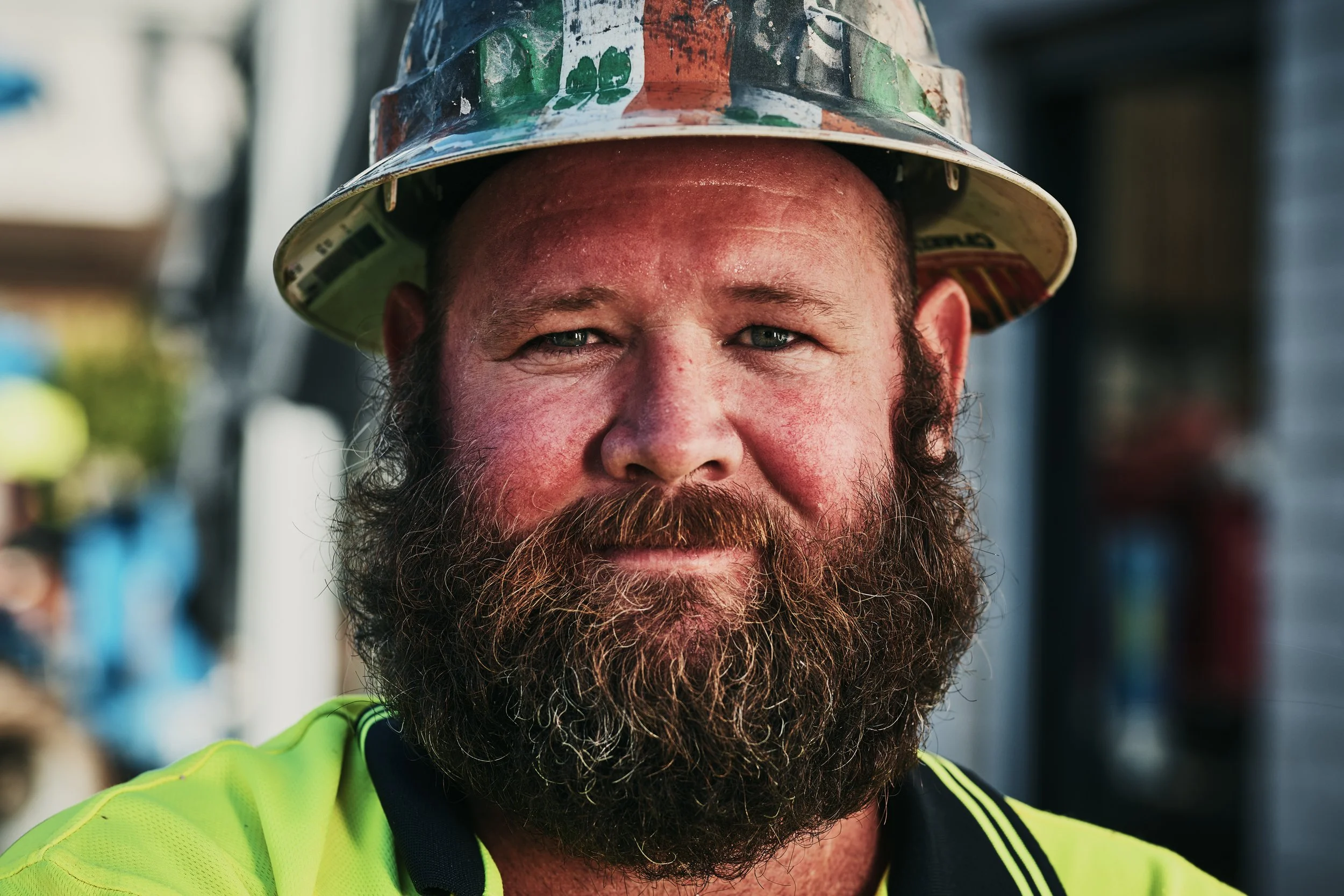 A man with a beard and short hair wearing a high-visibility yellow safety shirt and a colorful safety helmet, smiling outdoors on a clear day.