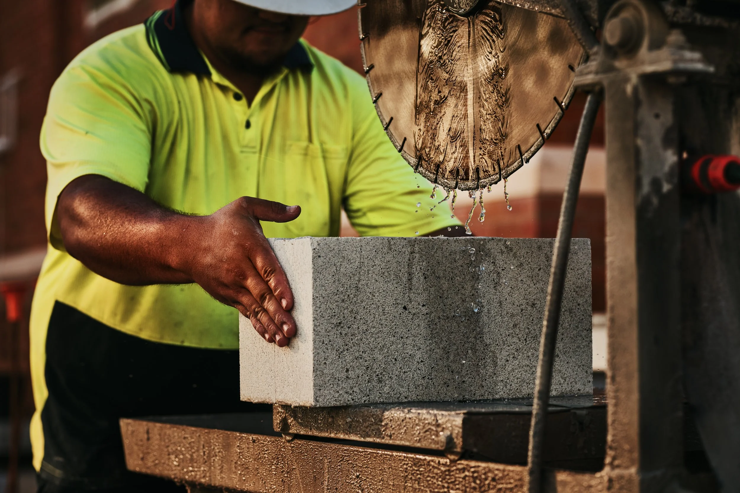 A man wearing a yellow safety vest is using a large tile saw to cut a piece of stone or concrete. Water is dripping from the saw as it cuts through the material.