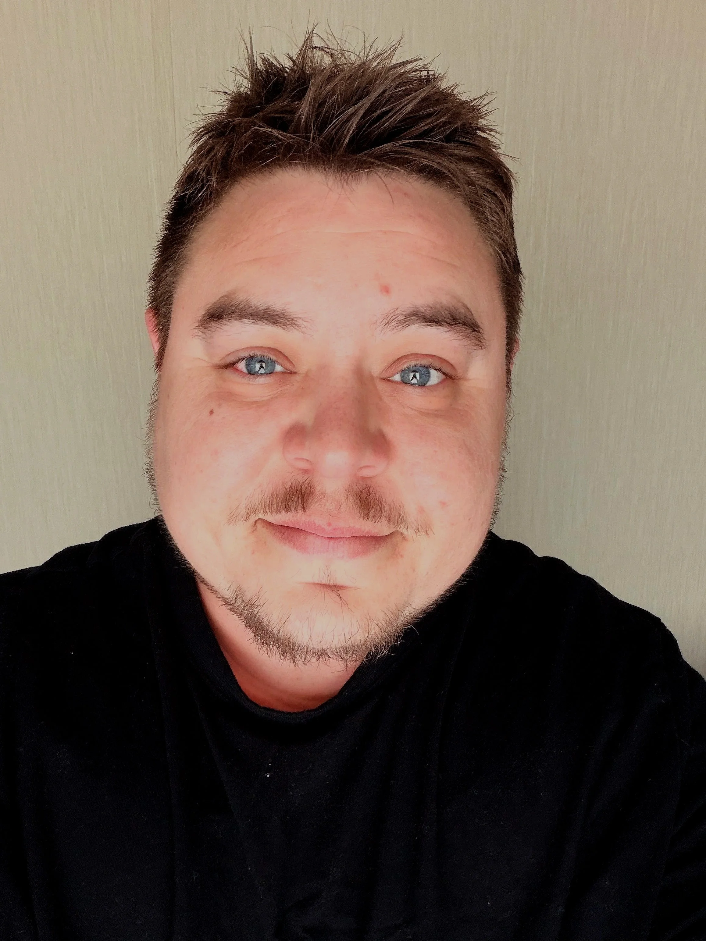 Self-portrait of a man with light blue eyes, light facial hair, and styled brown hair, wearing a black shirt, smiling slightly, against a plain beige background.