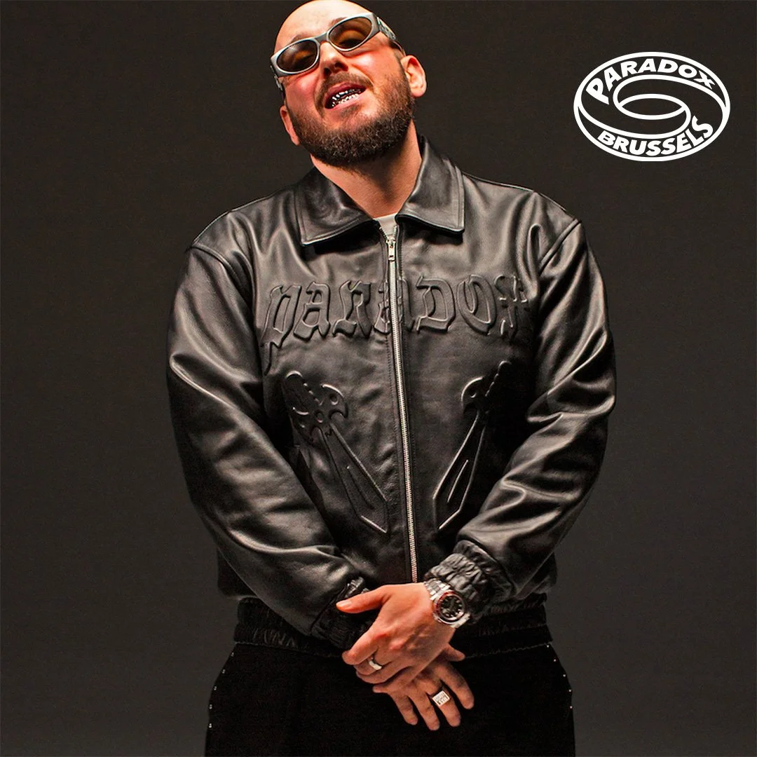 Un homme portant une veste en cuir noire, portant des lunettes de soleil, un collier en or, et plusieurs bagues, posant avec confiance contre un fond noir, avec un logo 'PARADOX BRSSELS' en haut à droite.
