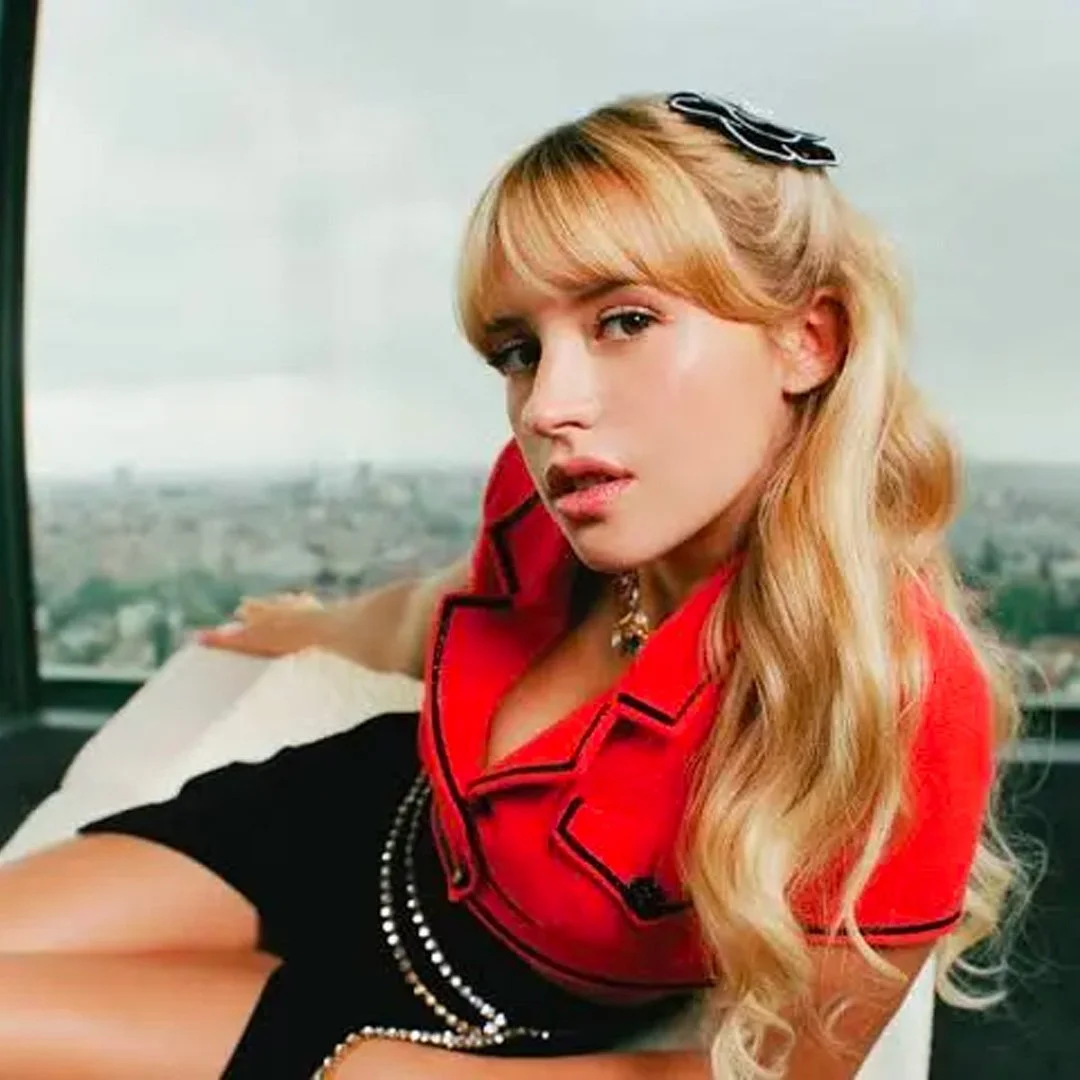 Jeune femme aux cheveux blonds ondulés, portant une veste rouge avec des détails noirs, assise près d'une fenêtre avec vue sur la ville.