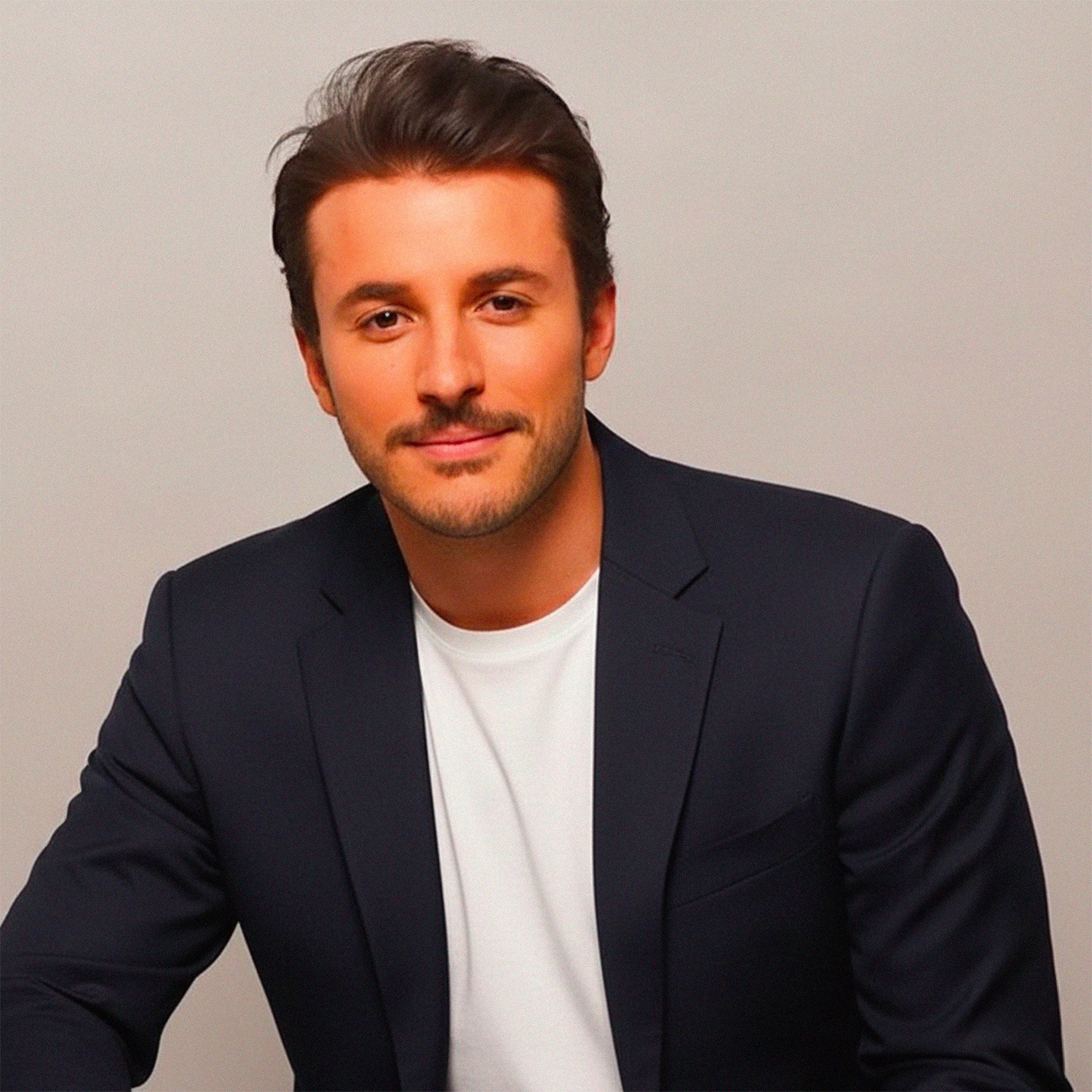 Jeune homme avec chaussette de cheveux bruns, portant un blazer noir et un T-shirt blanc, regardant en souriant vers l'avant, fond neutre