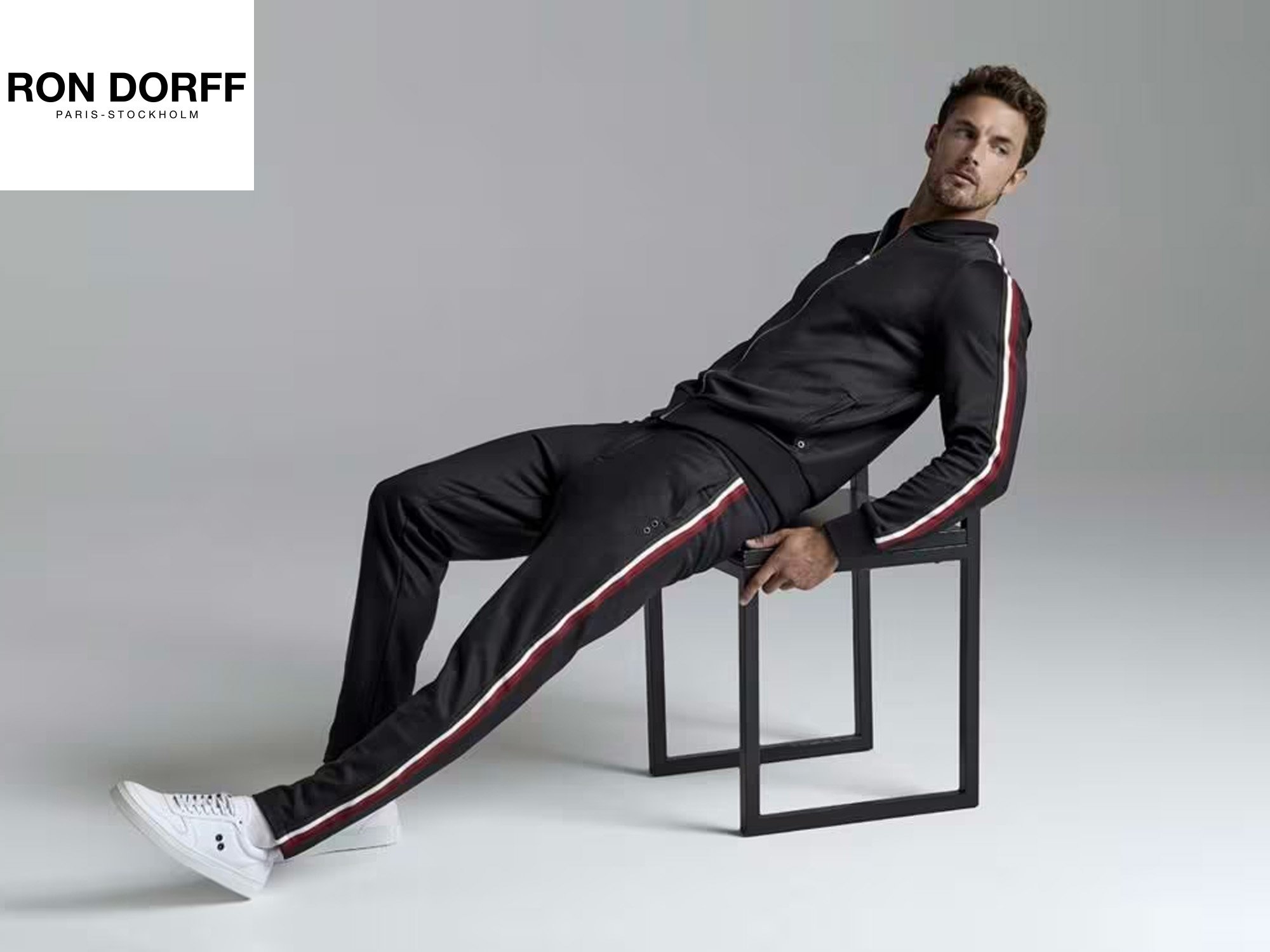Un homme portant une tenue de sport noire avec des bandes rouges et blanches, assis sur une chaise noire dans un studio avec fond gris.