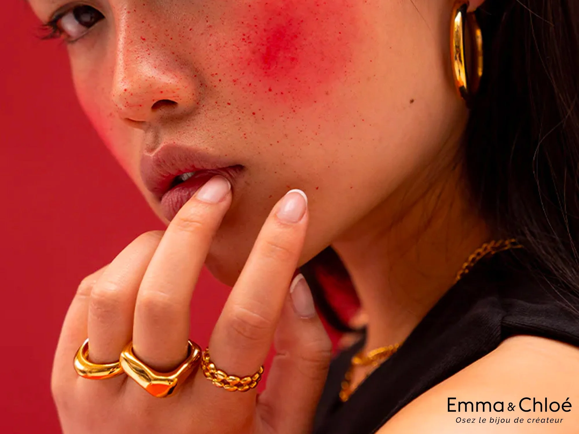 Zoom sur le visage d'une femme avec des taches de rousseur, portant des bagues en or, une boucle d'oreille en or, un col en chaîne, touchant ses lèvres avec ses doigts. Contexte rose.