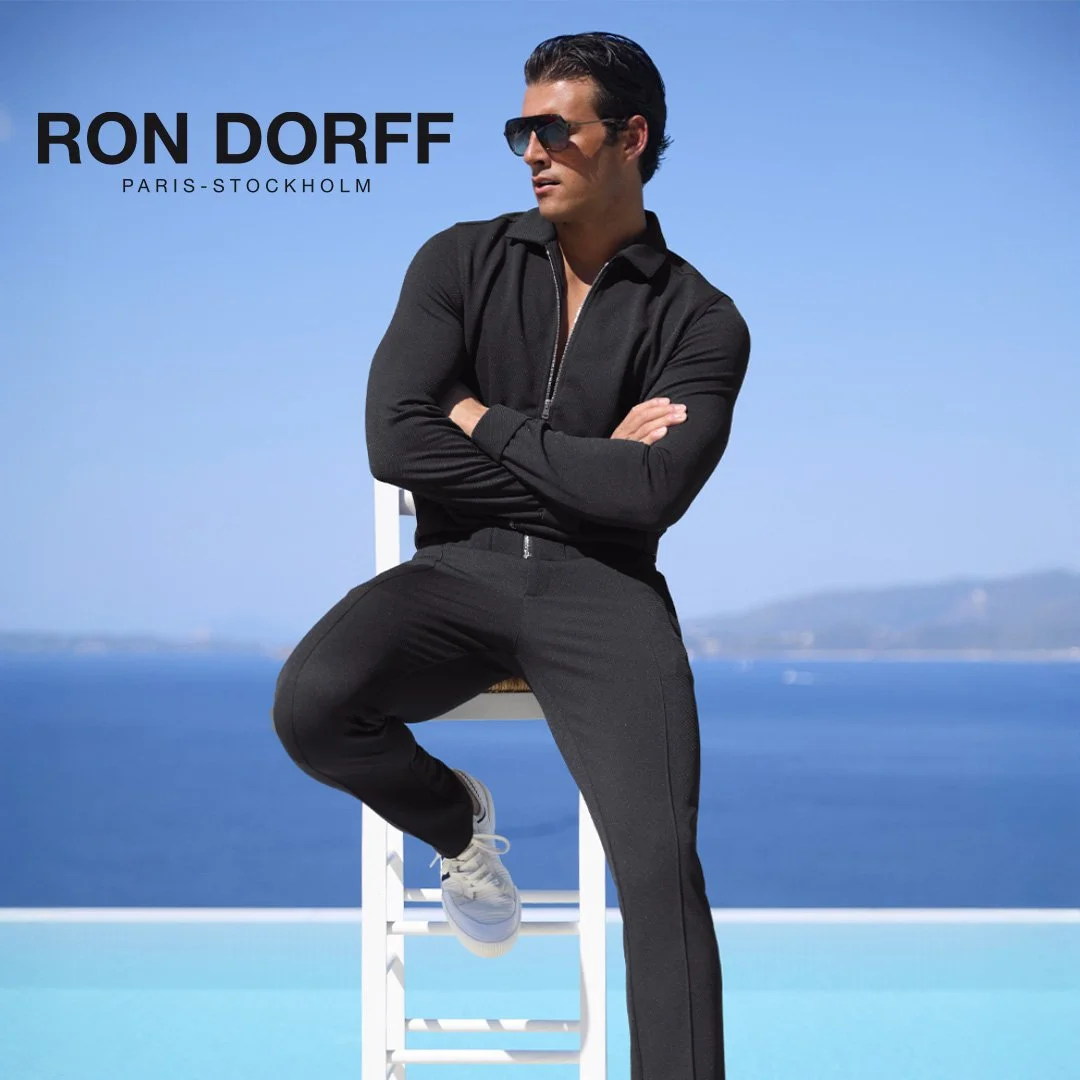 Un homme portant des vêtements noirs assis sur une chaise devant une mer bleue, portant des lunettes de soleil, avec le texte "Ron DorF PARIS-STOCKHOLM" dans le coin supérieur gauche.