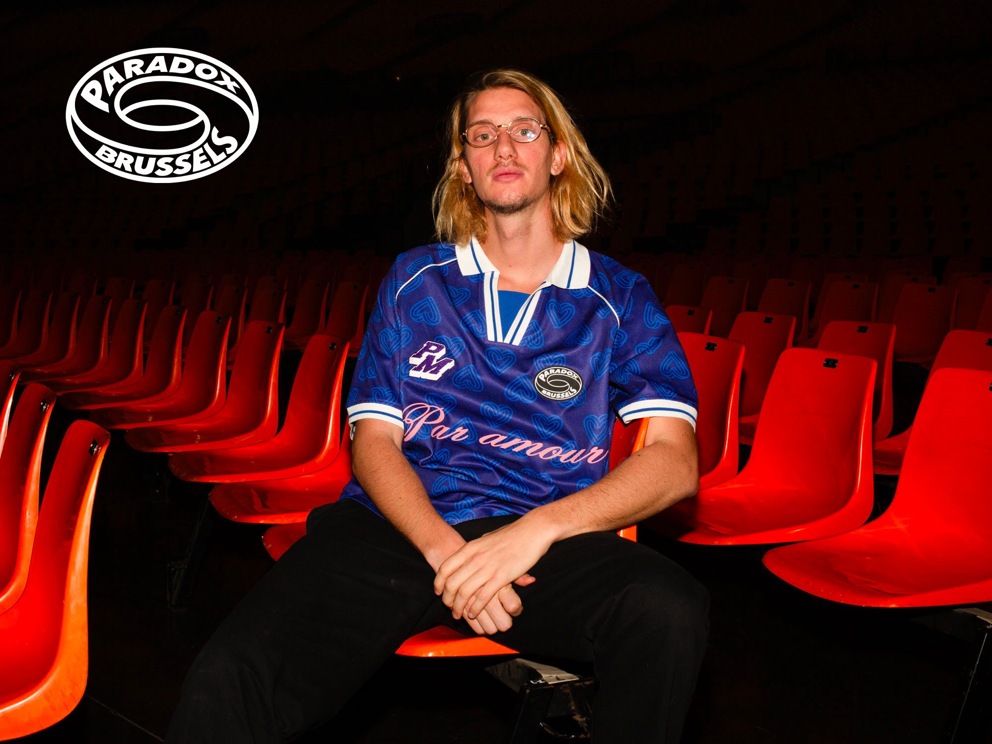 Un homme assis sur des sièges rouges dans une salle sombre, portant un maillot de hockey bleu avec des motifs en cœur, et un logo