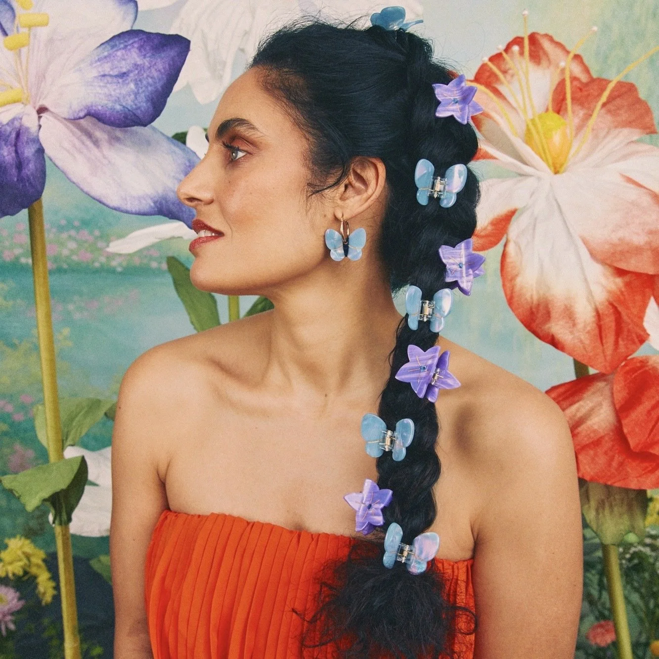 Une femme avec de longs cheveux noirs tressés décorés de papillons en plastique bleu et violet, portant une boucle d'oreille papillon bleue, pose devant un fond coloré de fleurs gigantesques.