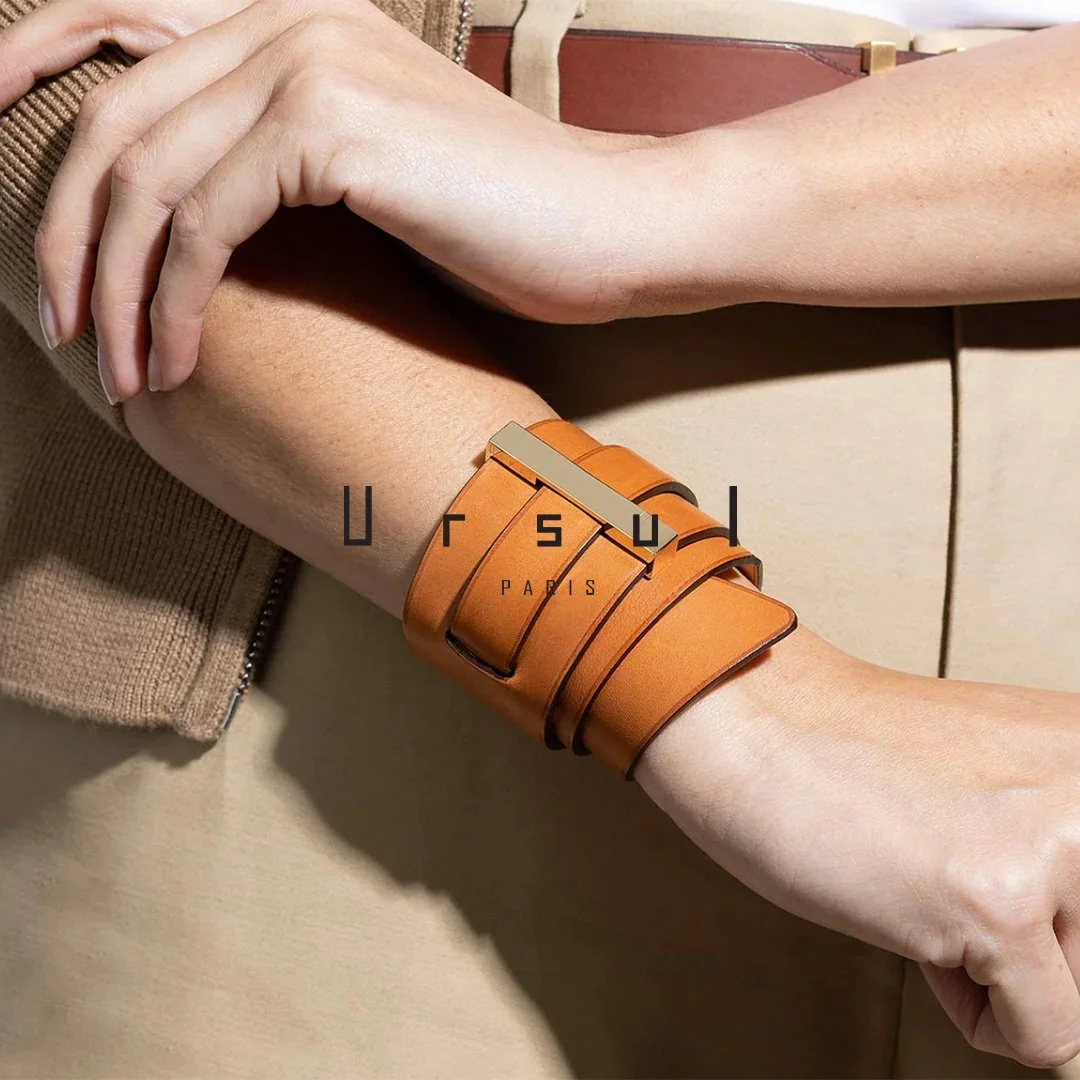 Un bras portant un bracelet en cuir orange avec une plaque métallique, le bras repose sur une autre main. Fond beige, logo « Oursu Paris » en superposition.