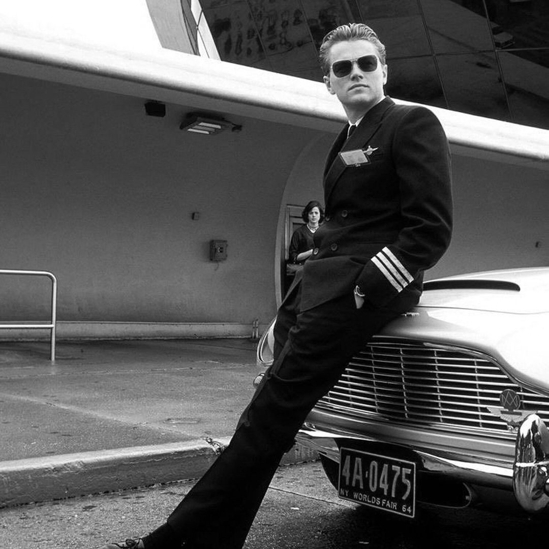 Un homme portant un costume d'officier de l'aviation de ligne, avec des lunettes de soleil, pose contre une voiture de style vintage, dans un décor urbain moderne.