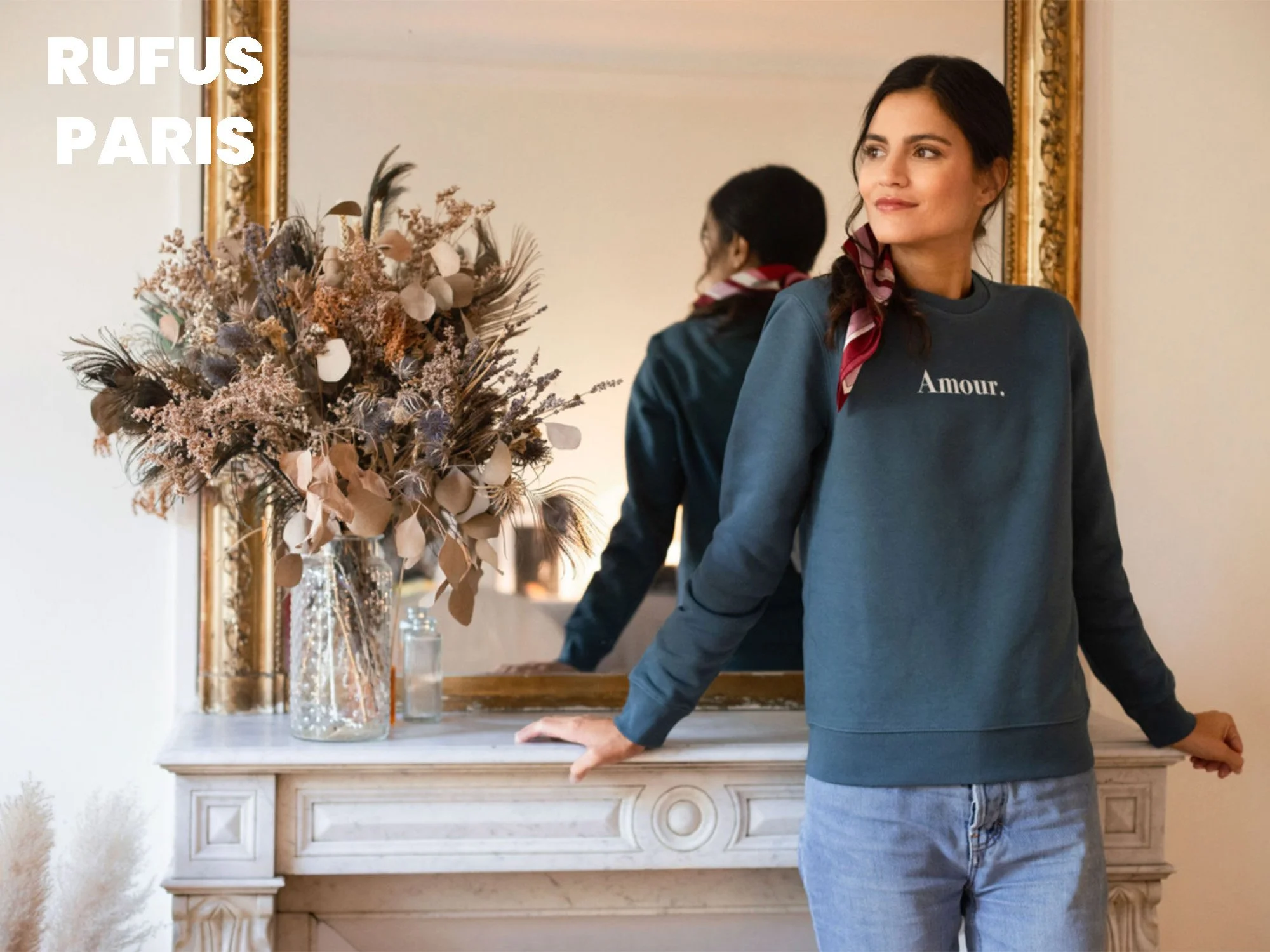 Une femme portant un sweat bleu avec l'inscription 'Amour.', se tenant devant un miroir dans une pièce élégante, avec un bouquet de fleurs séchées dans un vase en verre sur une cheminée en marbre.