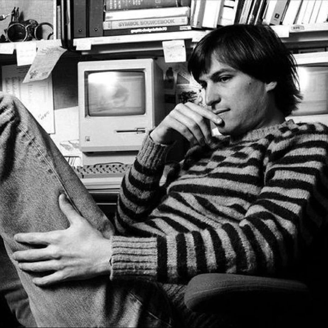 Un homme assis dans une pièce remplie de livres et de vieux équipements, portant un pull rayé, appuyé sur sa main, regardant pensivement.