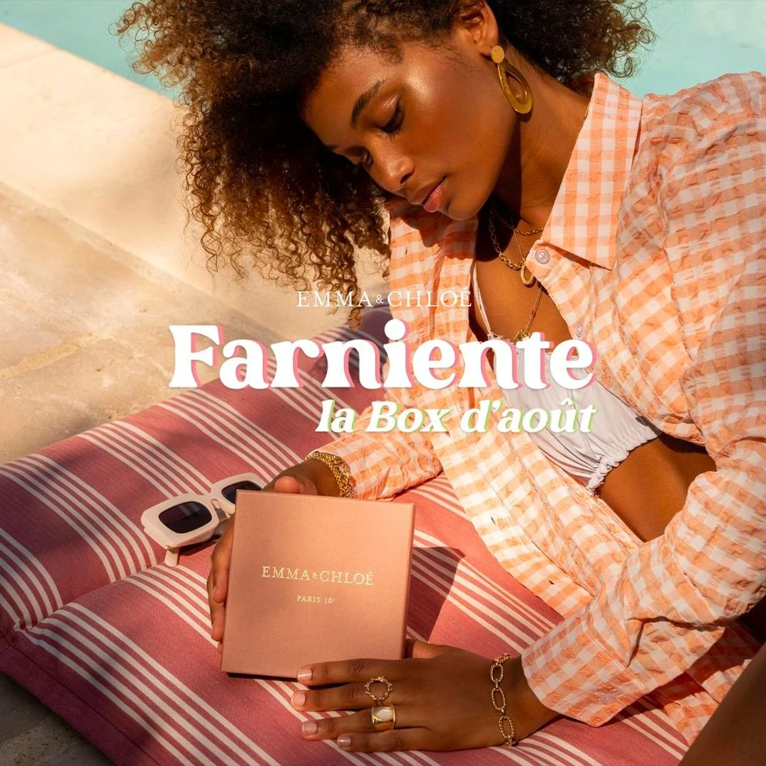 Une femme souriante portant une chemise à carreaux orange et blanc, avec des bijoux dorés, regarde un coffret de bijoux Emma & Chloé, posé sur un coussin rayé. La scène est en plein air, avec une lumière chaleureuse.