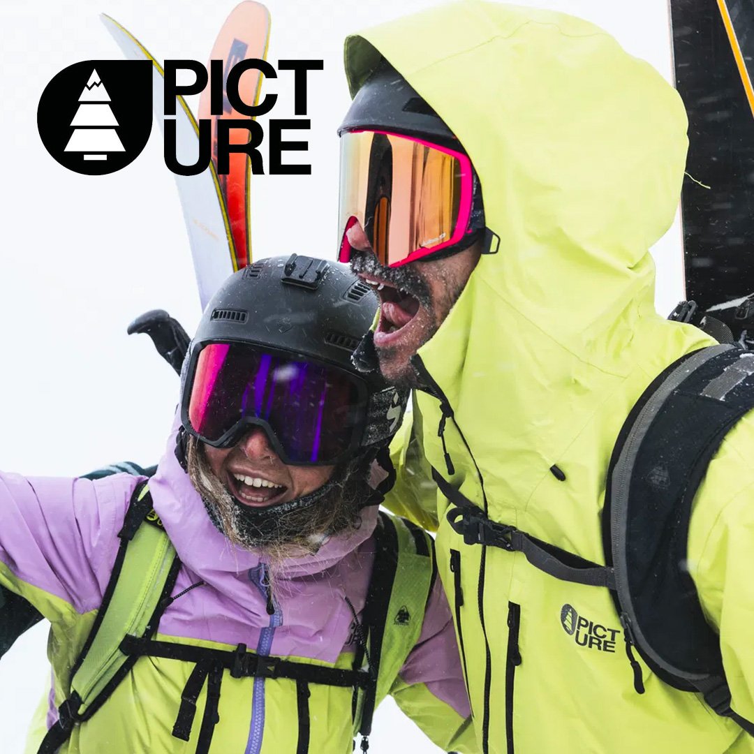 Deux personnes en équipement de ski enneigé, souriantes, posant pour une photo de selfie en bas de la montagne, portant des vestes jaunes et violettes, avec casques et lunettes de ski.