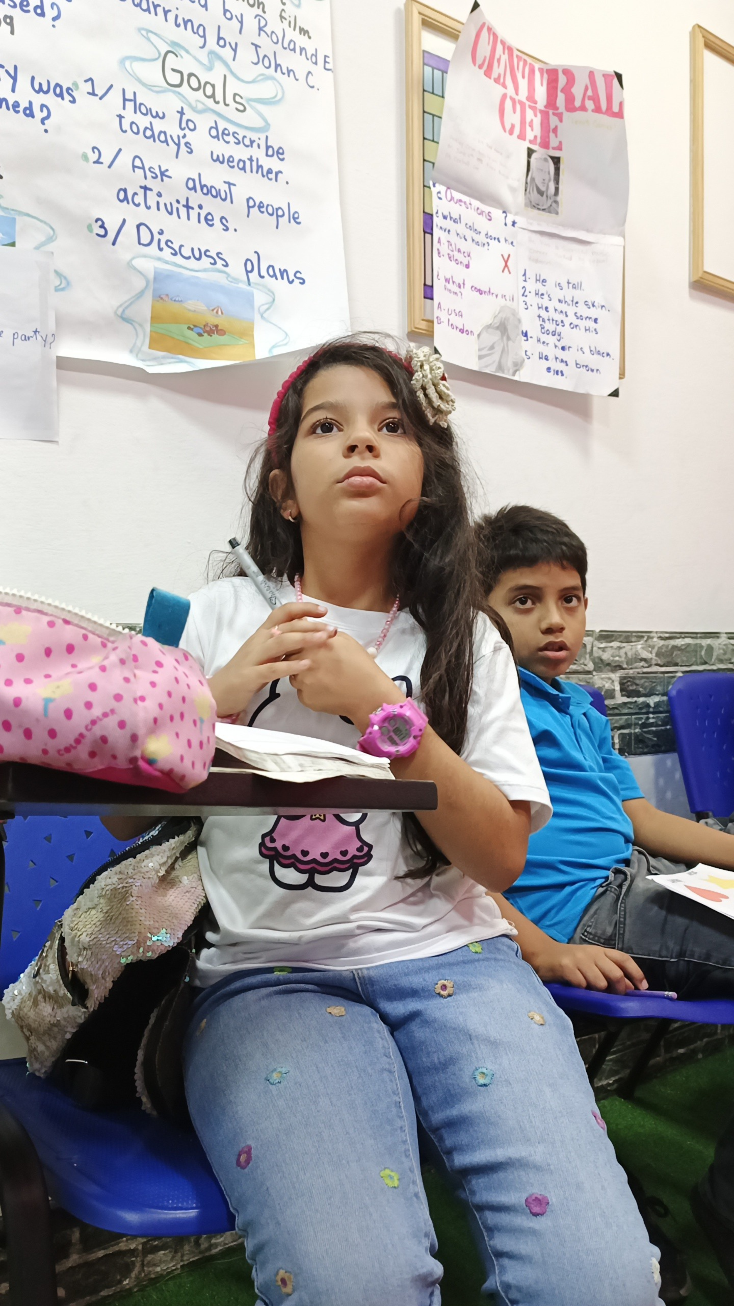 Niña con camiseta blanca con un dibujo de una cupcake, pantalones azules con bordados de flores, y una pulsera rosa, sentado en una silla azul con estudiantes en un aula, con carteles en la pared con notas y dibujos.