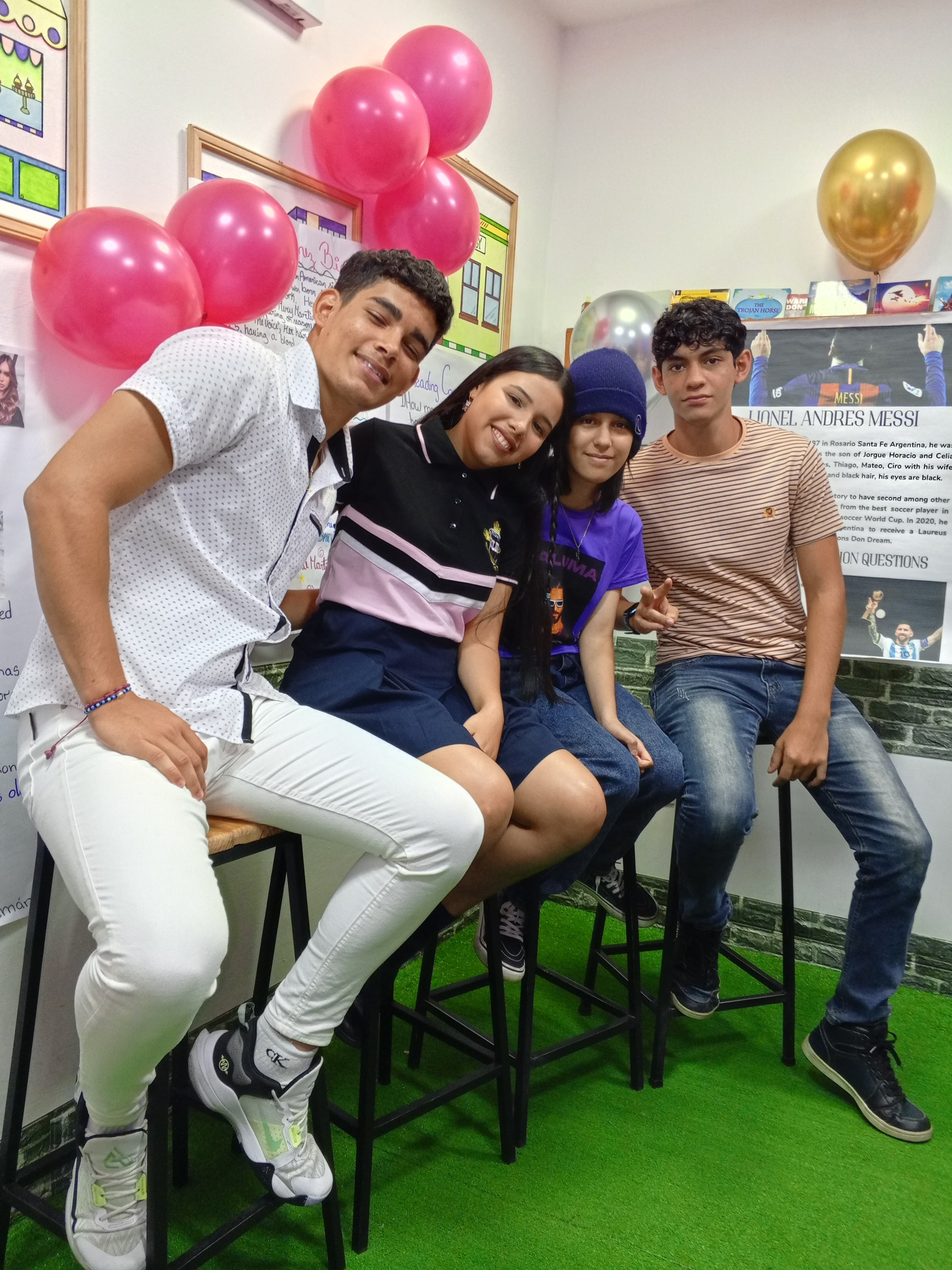 Cuatro jóvenes sentados sobre taburetes en una sala decorada con globos rosas, dorados y plateados, y posters en las paredes. Están sonriendo y posando para la foto.