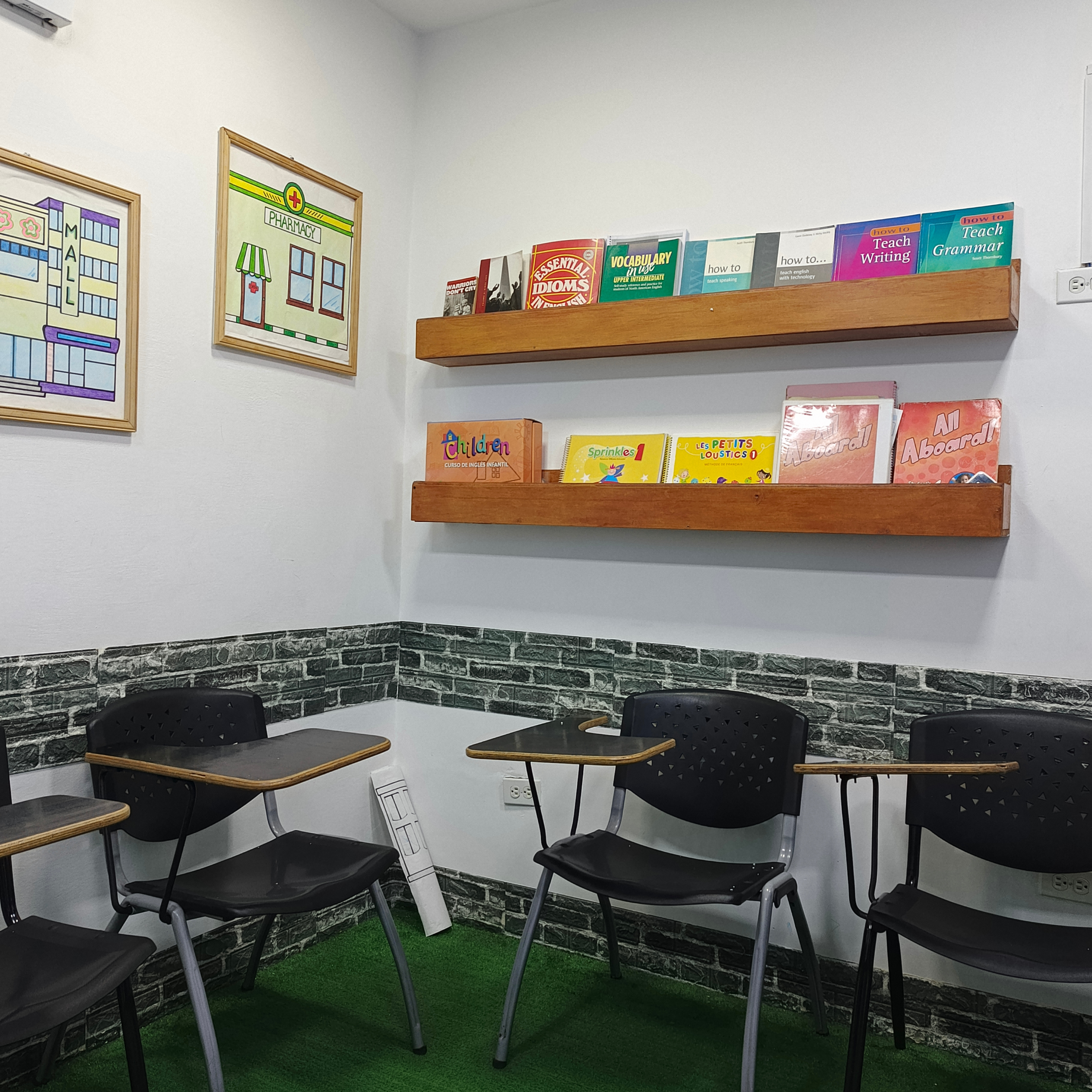 Sala de clases con sillas negras y escritorios de madera, paredes blancas y decoraciones con libros y pósters educativos.