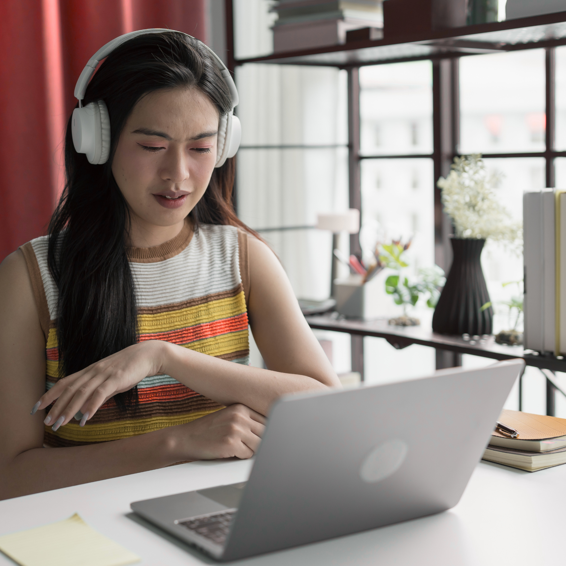 Joven mujer con auriculares en una oficina mirando la pantalla de una computadora portátil, con plantas y objetos decorativos en el fondo.
