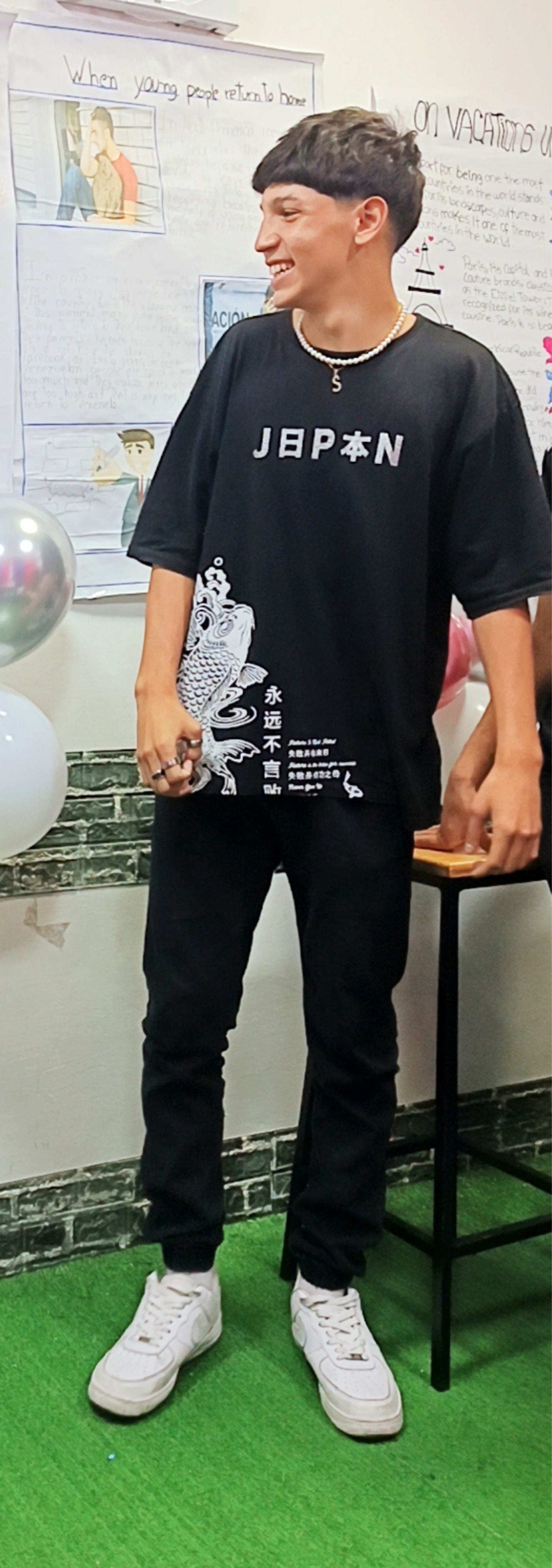 Joven hombre sonriendo, usando una camiseta negra con diseño de pez y palabras en japonés y chino, pantalones negros y zapatillas blancas, en un ambiente de aula con carteles en la pizarra y globos blancos.
