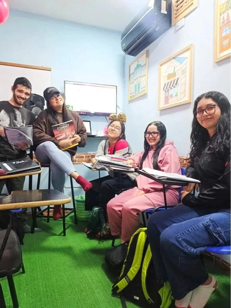 Grupo de cinco jóvenes en una sala de estudio, algunos con cuadernos y libros, sonriendo y posando para la foto, con paredes decoradas y un aire acondicionado en la esquina.