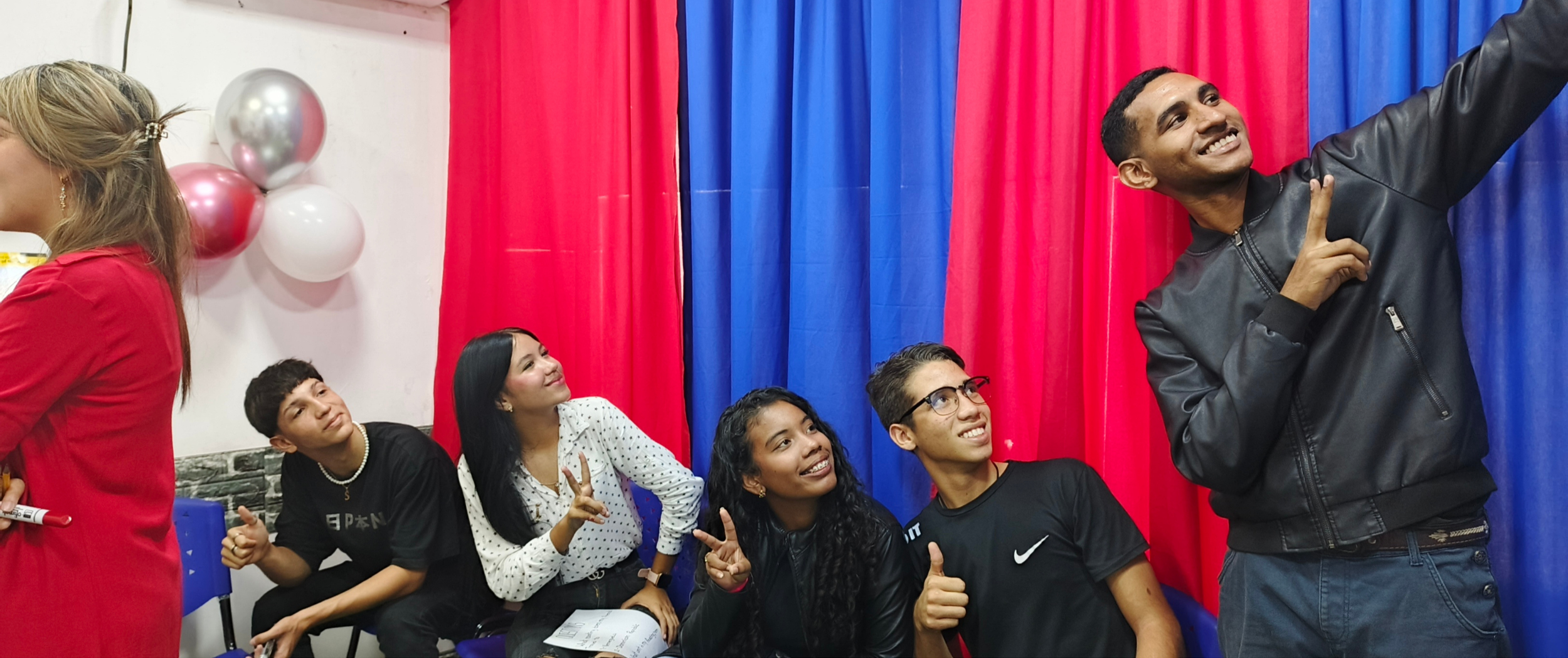 Un grupo de personas jóvenes tomando una selfie en un evento con fondo de cortinas rojas y azules.