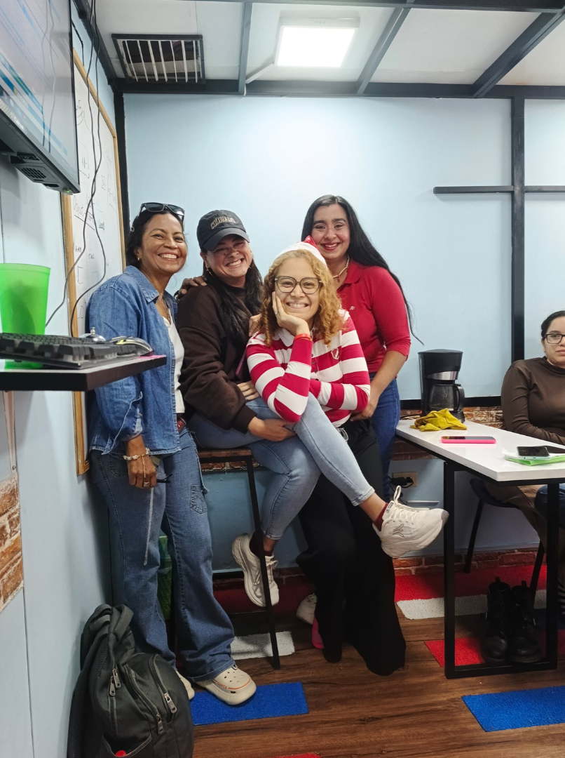 Cuatro mujeres sonriendo y posando en una sala de clases o reunión, una está sentada en una silla, las otras tres paradas a su lado.