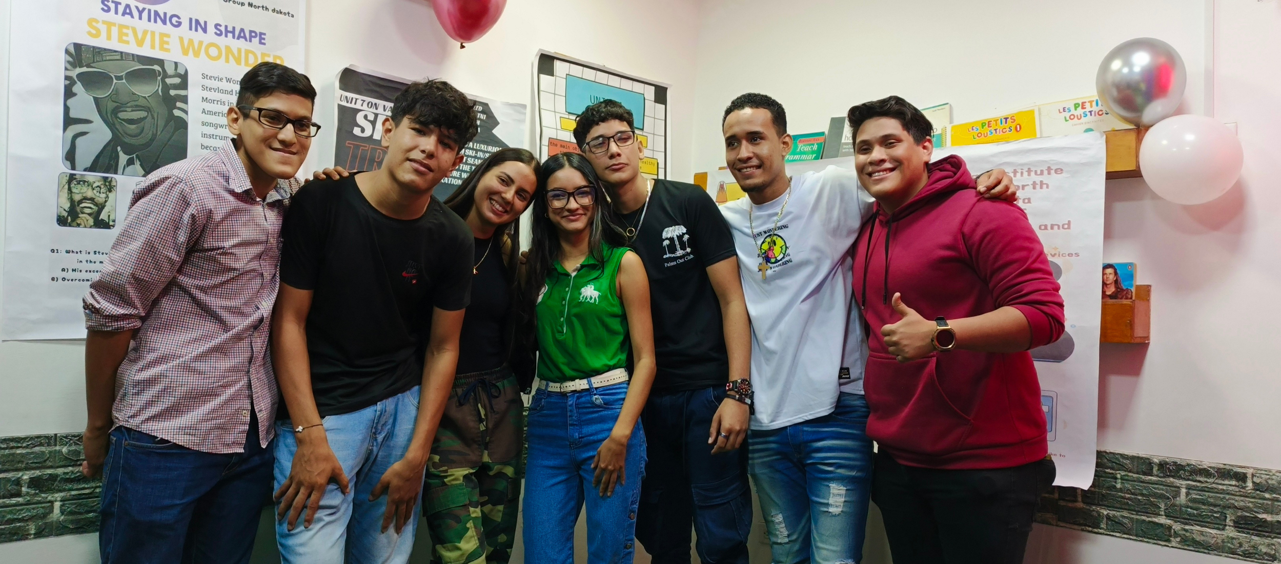 Grupo de siete jóvenes sonriendo y posando juntos en un aula con posters y globos en el fondo.