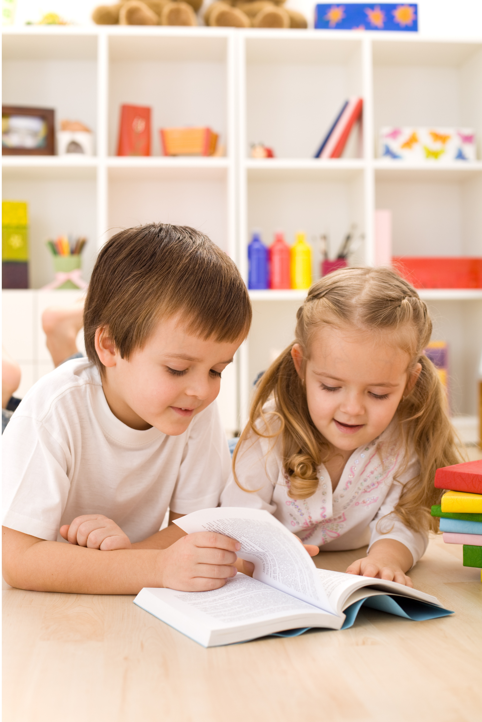 Dos niños, un niño con cabello castaño y una niña con cabello rubio y trenzado, están leyendo un libro juntos sobre una mesa de madera en un aula. Al fondo, hay estanterías llenas de libros, papelería y materiales de arte en un aula escolar.