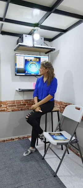 Mujer sentada en una sala de reuniones con una pantalla y un proyector en la pared, sosteniendo una carpeta, en un ambiente profesional.