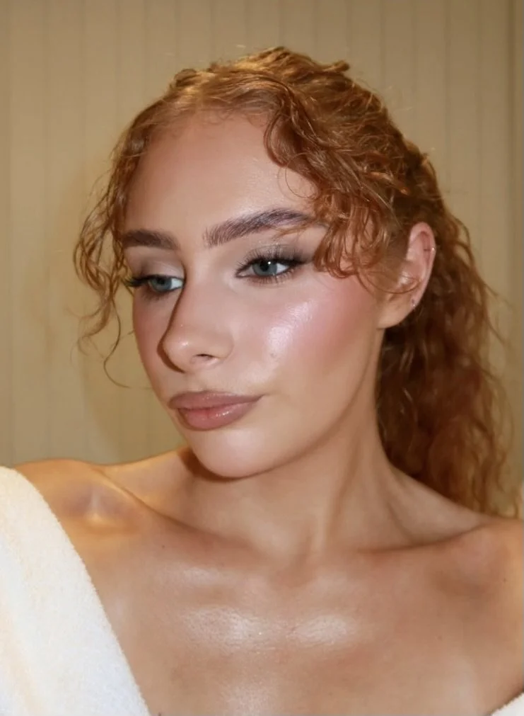 Close-up of a woman with curly red hair, blue eyes, and makeup, wearing a white towel on her shoulder.
