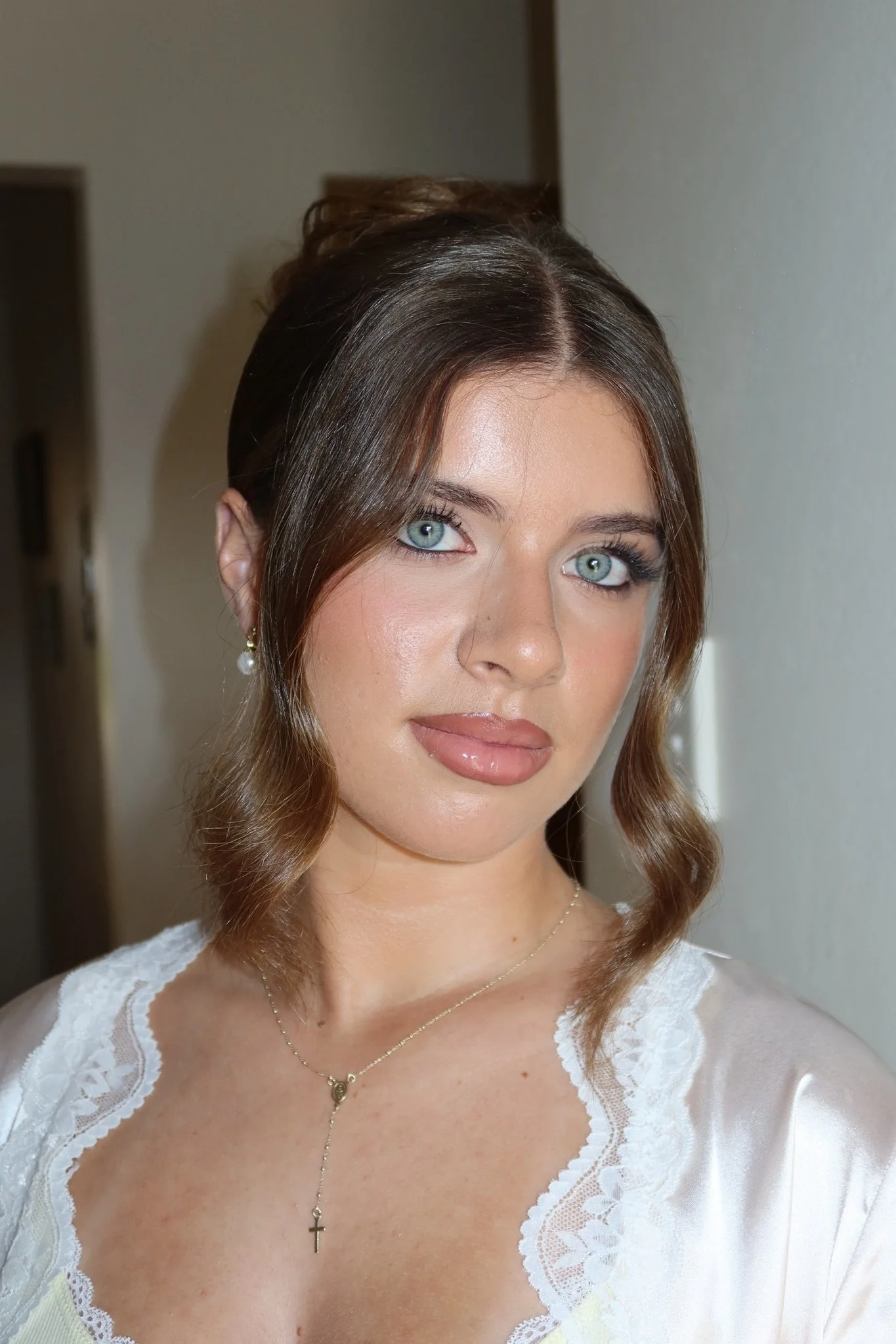 A woman with wavy brown hair, blue eyes, light makeup, wearing pearl earrings, a gold necklace with a cross pendant, and a cream-colored top with lace details.